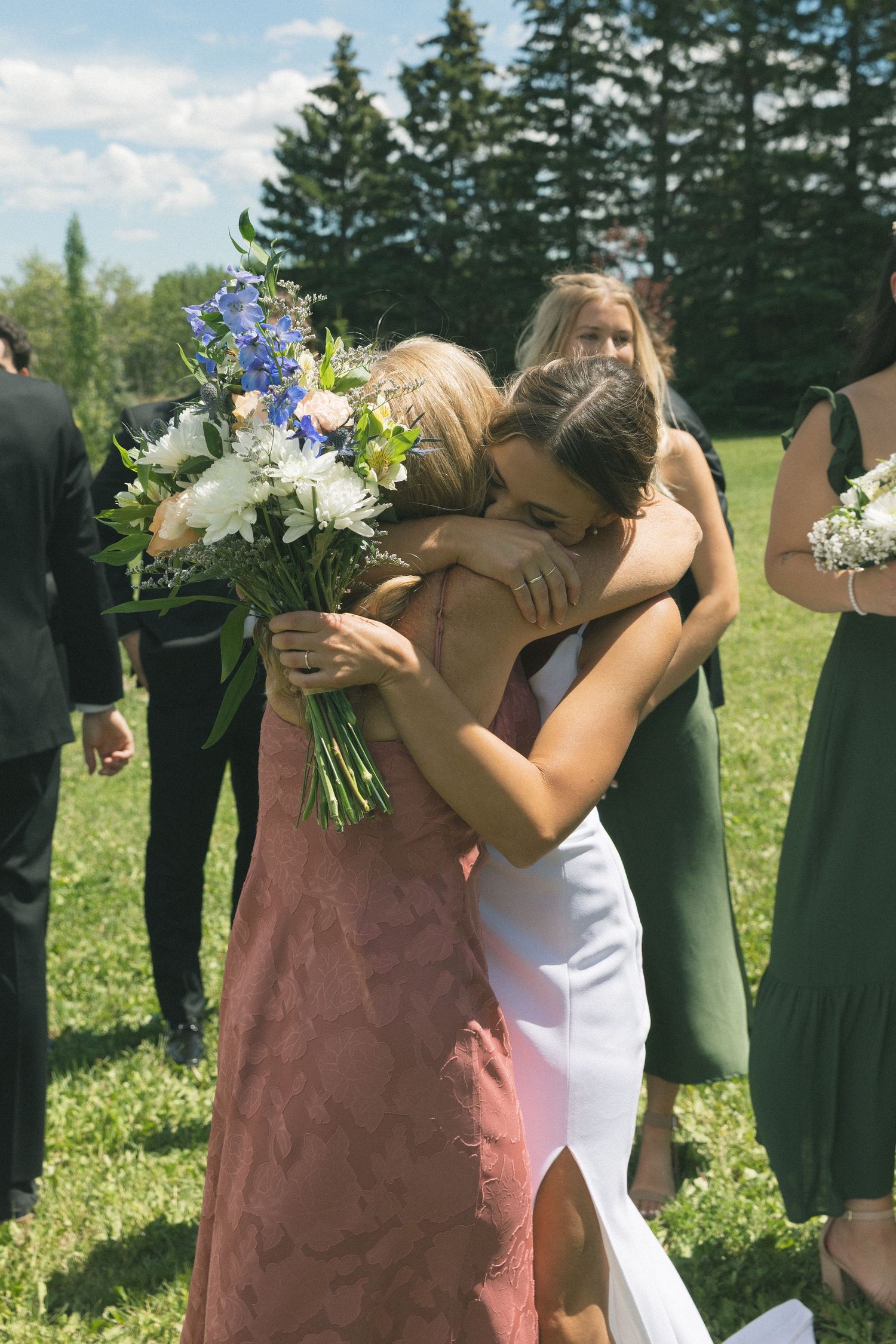 Bride in white dress embraces guest warmly while holding blue and white floral bouquet at outdoor wedding reception.