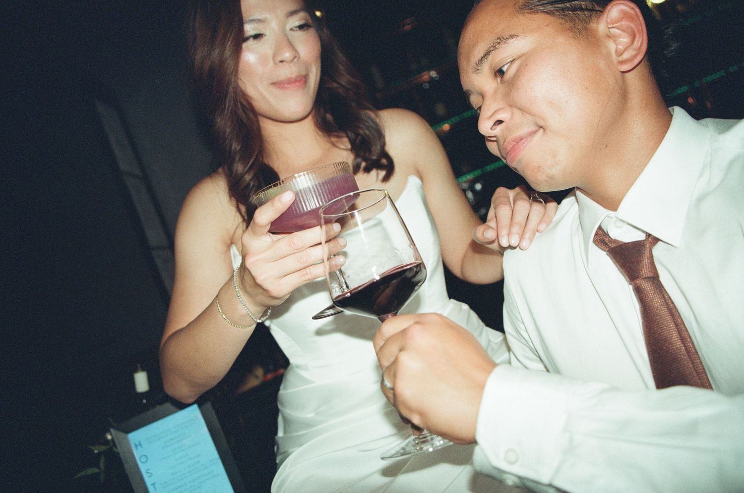 A woman in white dress and man in white shirt share red wine glasses at a social event.