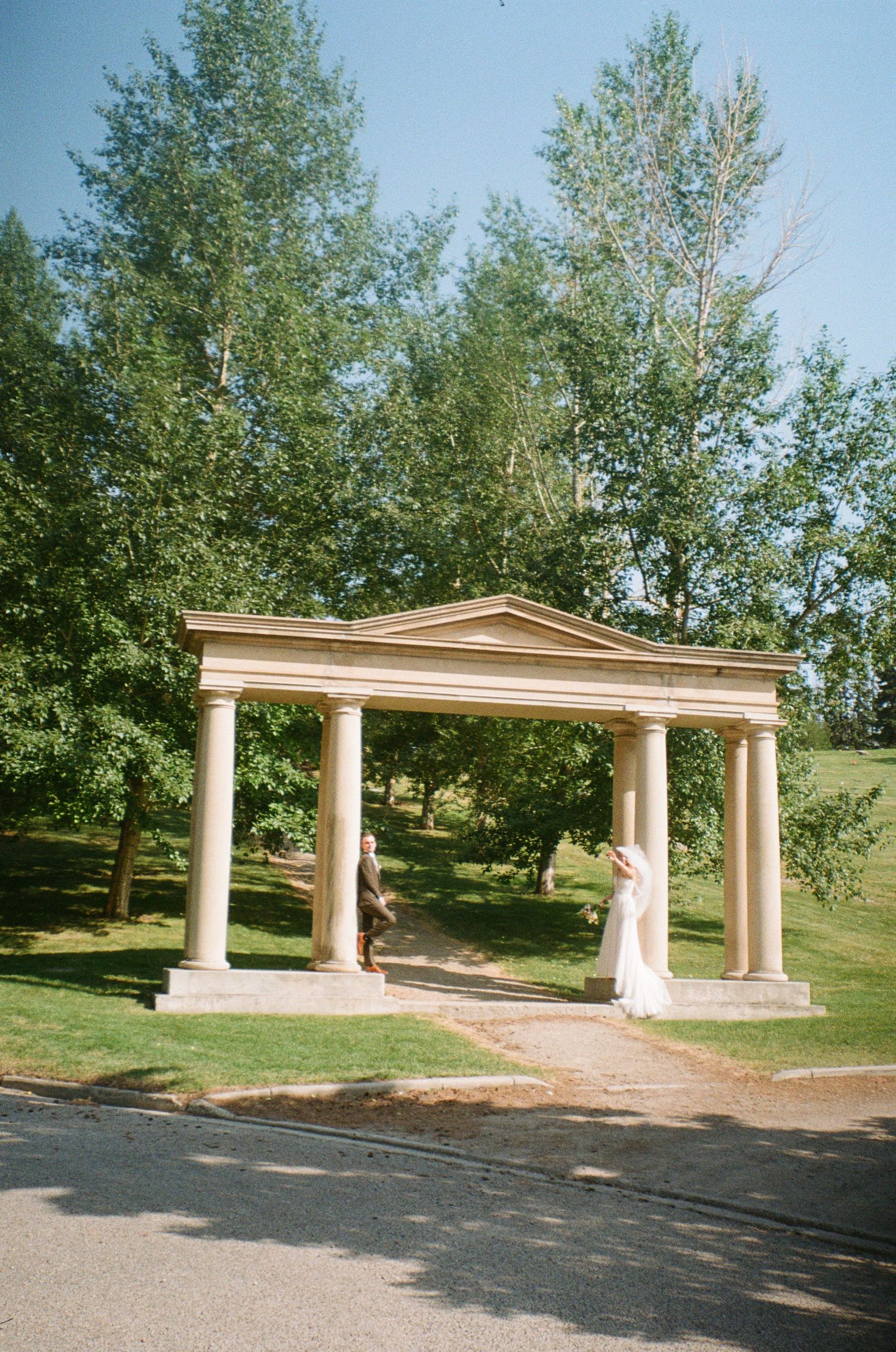 A classical stone pergola with white columns and triangular pediment stands among green trees in a park setting.