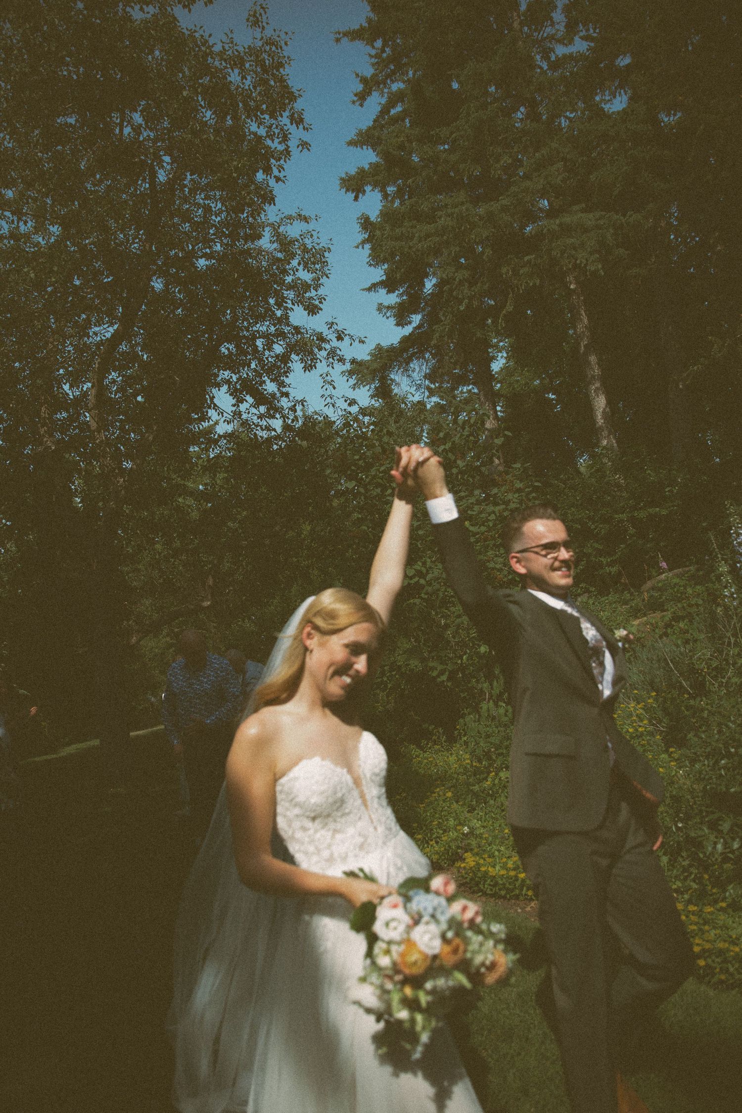 A joyful wedding celebration outdoors with a couple in formal attire holding hands against a backdrop of green trees.