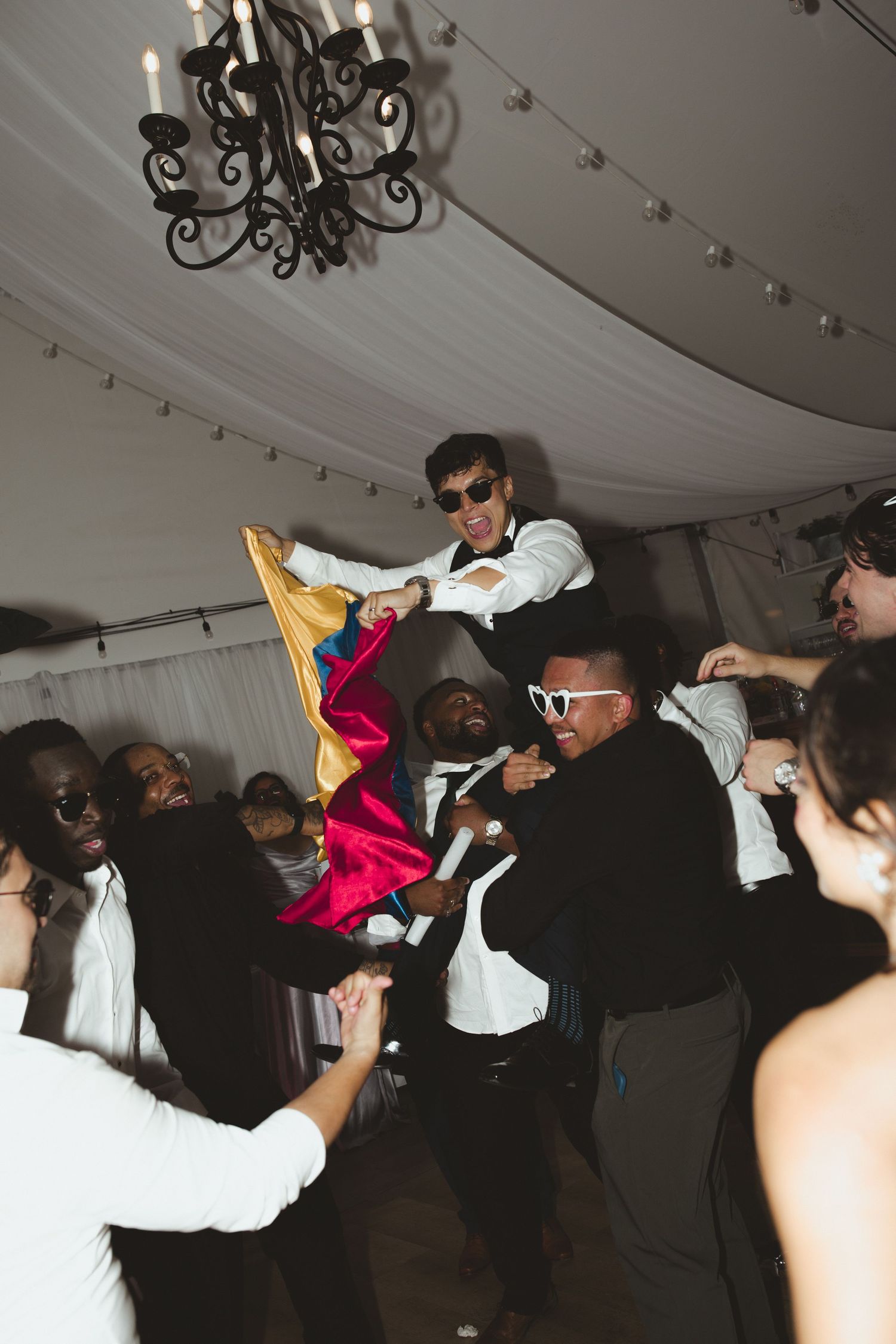 Groomsmen lifting groom at wedding reception party under chandelier in tent venue.