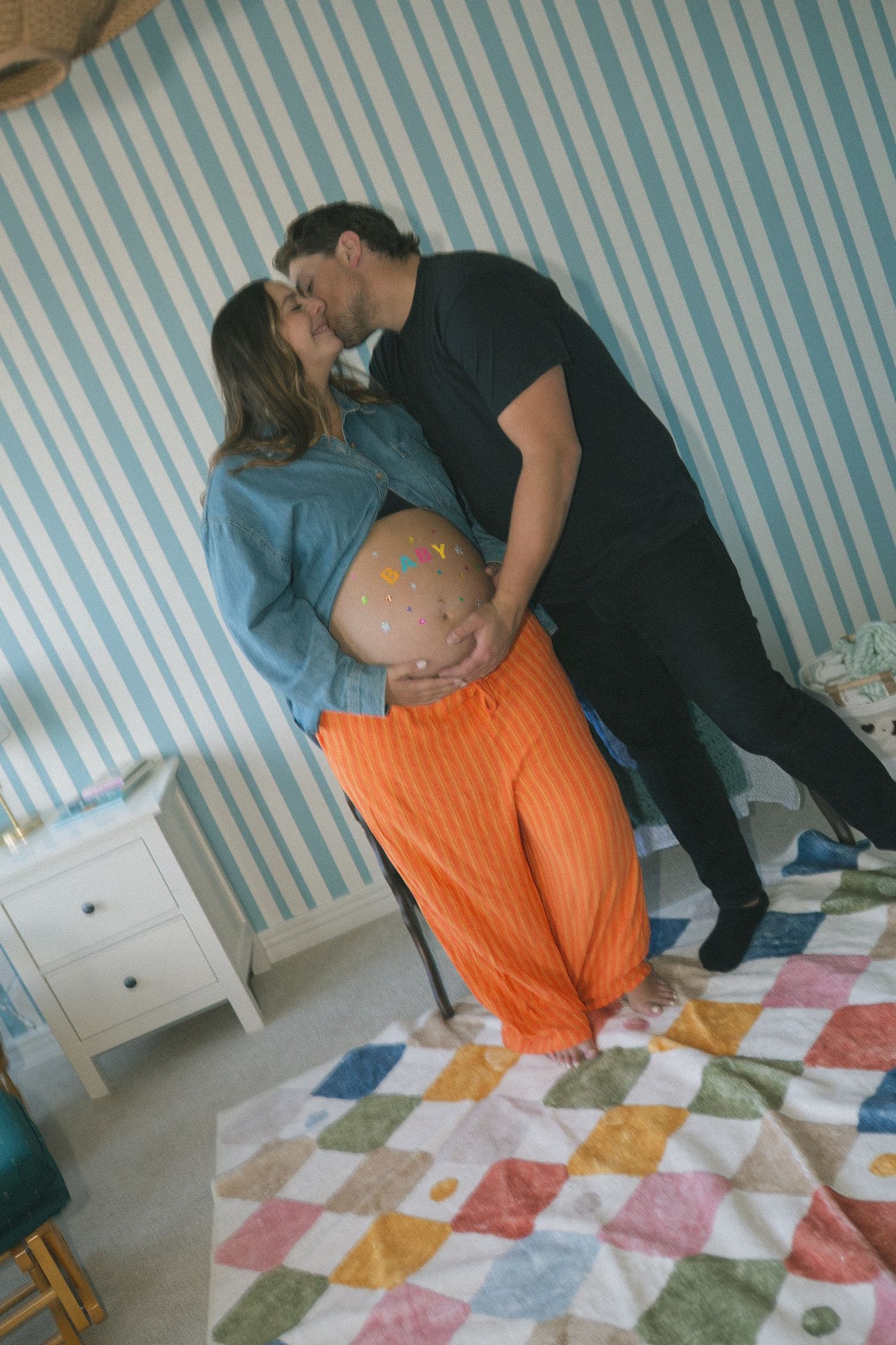 A couple embraces in a nursery with blue striped wallpaper and a colorful patchwork rug.