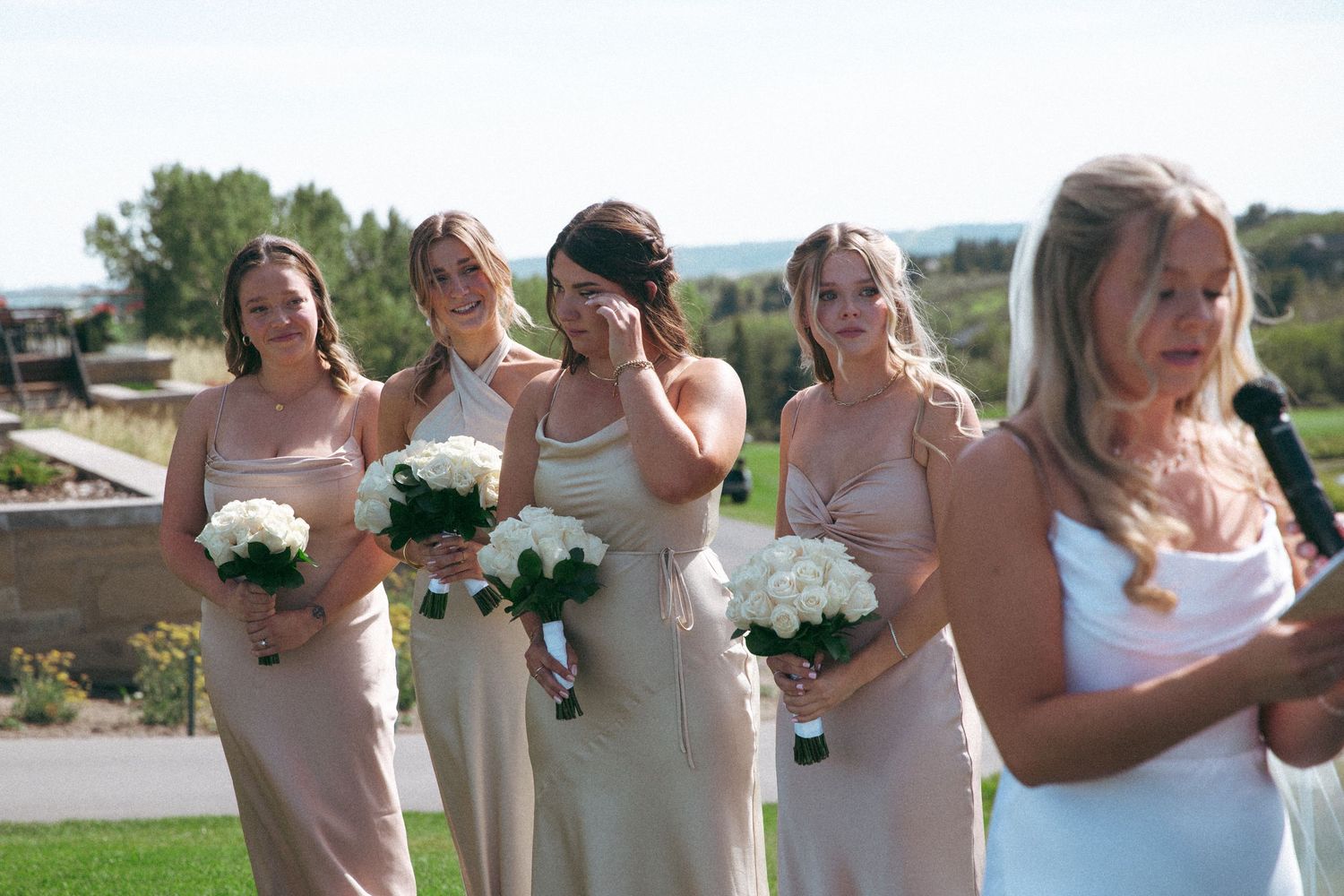 Bridesmaids in neutral dresses holding white bouquets during outdoor ceremony.
