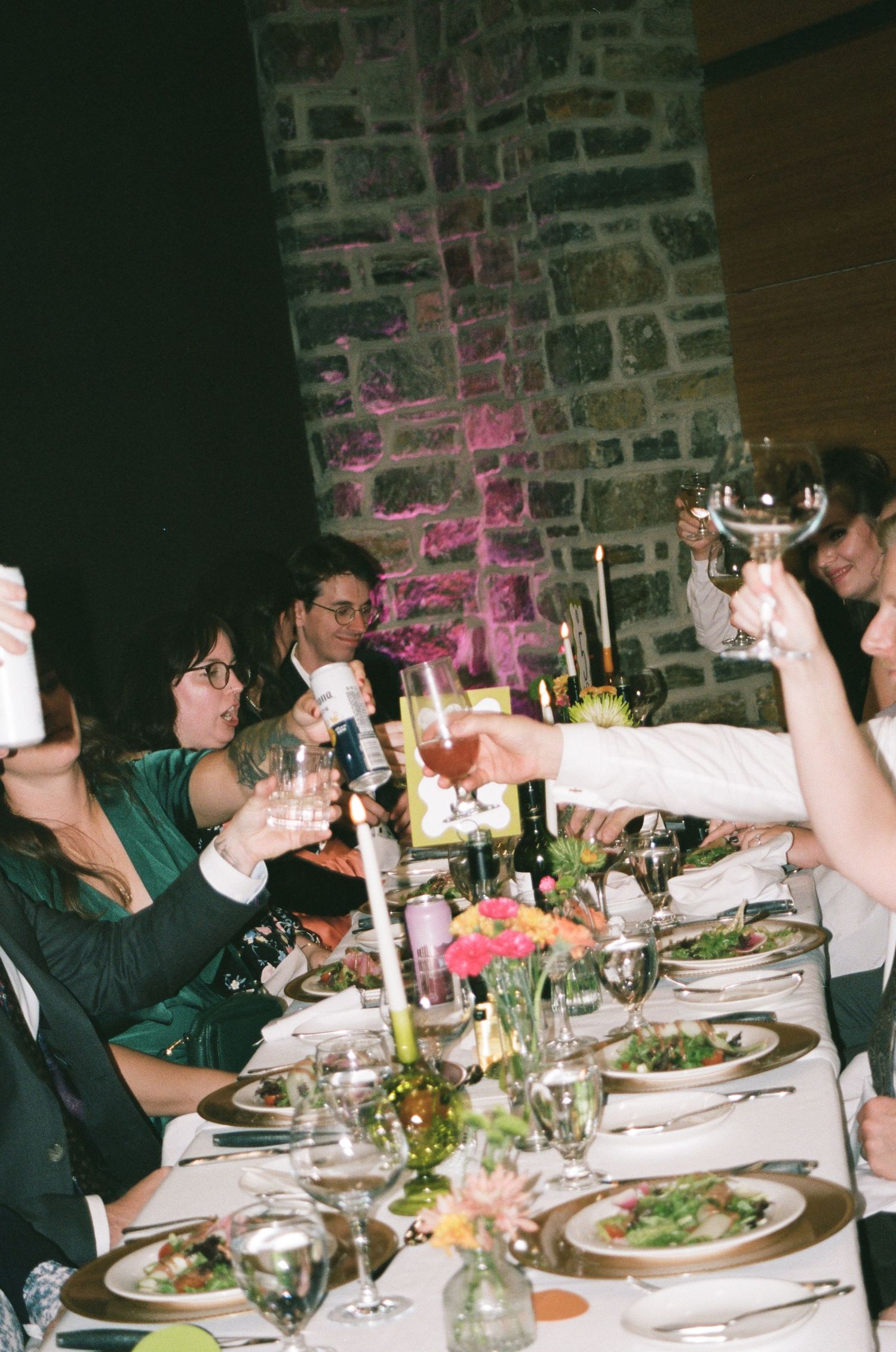Guests raising glasses in a toast at an elegant wedding reception dinner with floral centerpieces and candlelight.