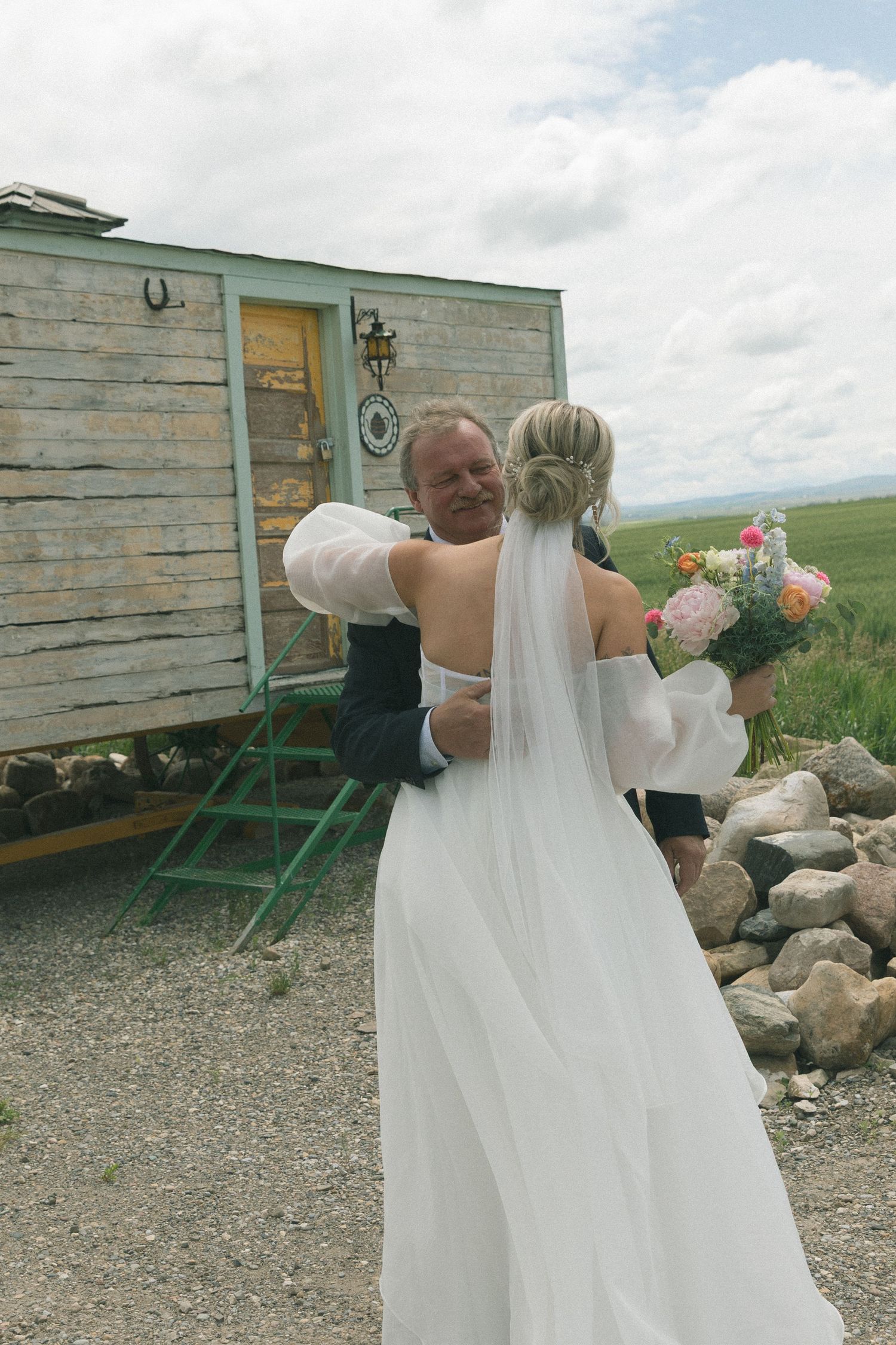 A bride in a flowing white dress poses near a rustic wooden building in a rural setting.