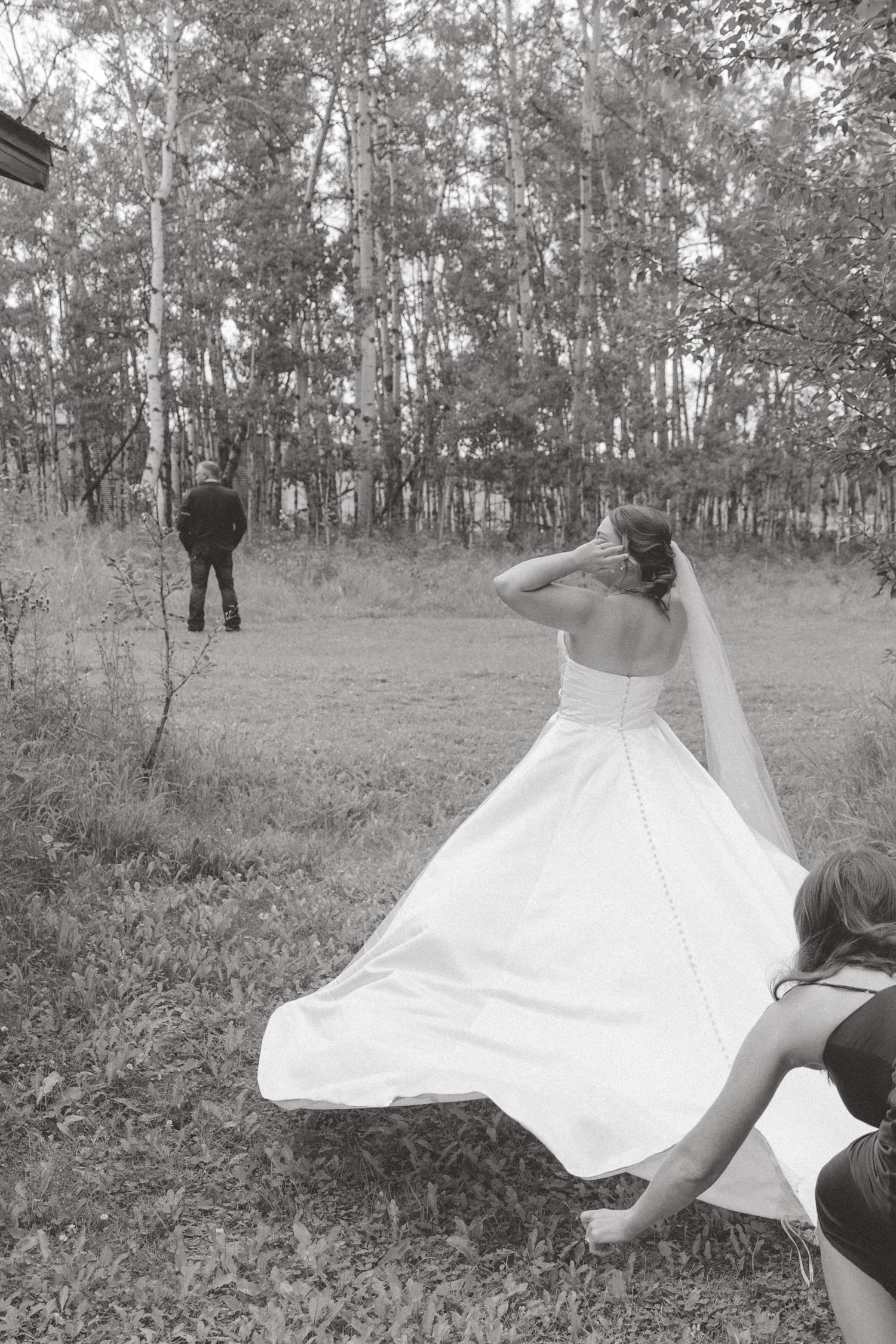 Black and white photo of a bride in a flowing white gown and veil walking toward a groom in a forest clearing.