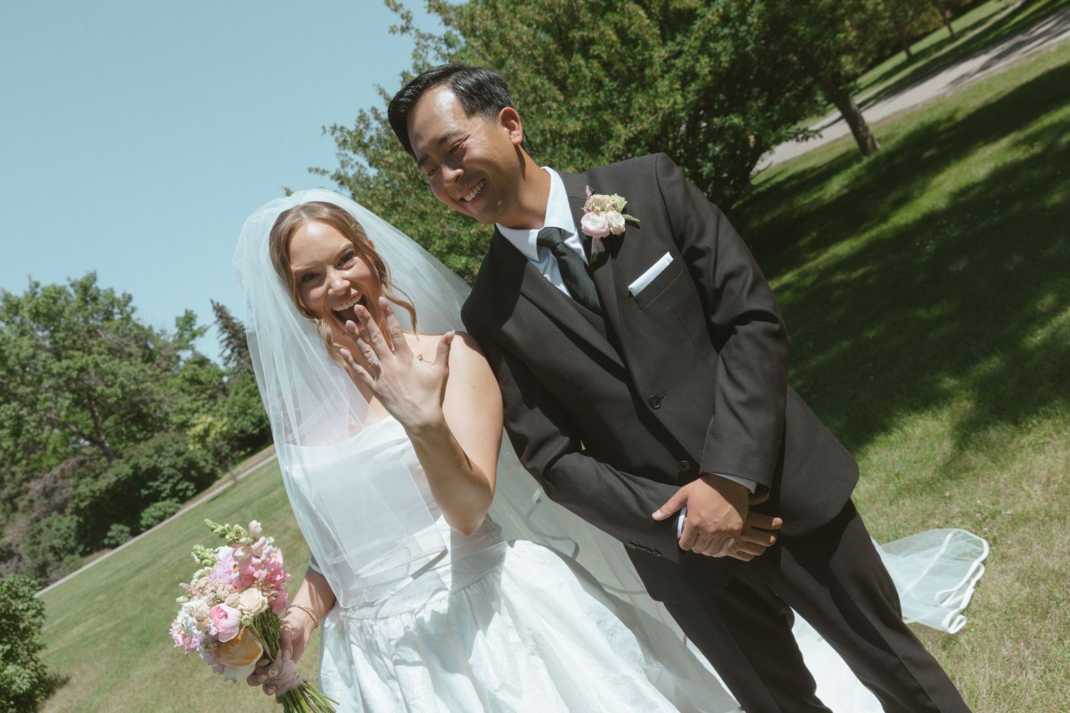 Happy newlywed couple posing outdoors on sunny day, bride in white gown with veil holding pink bouquet, groom in black suit.