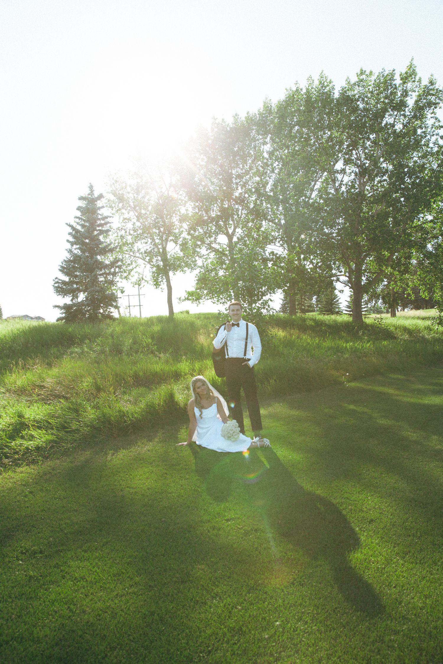 A couple sits on a grassy hillside in wedding attire as sunlight streams through the trees.