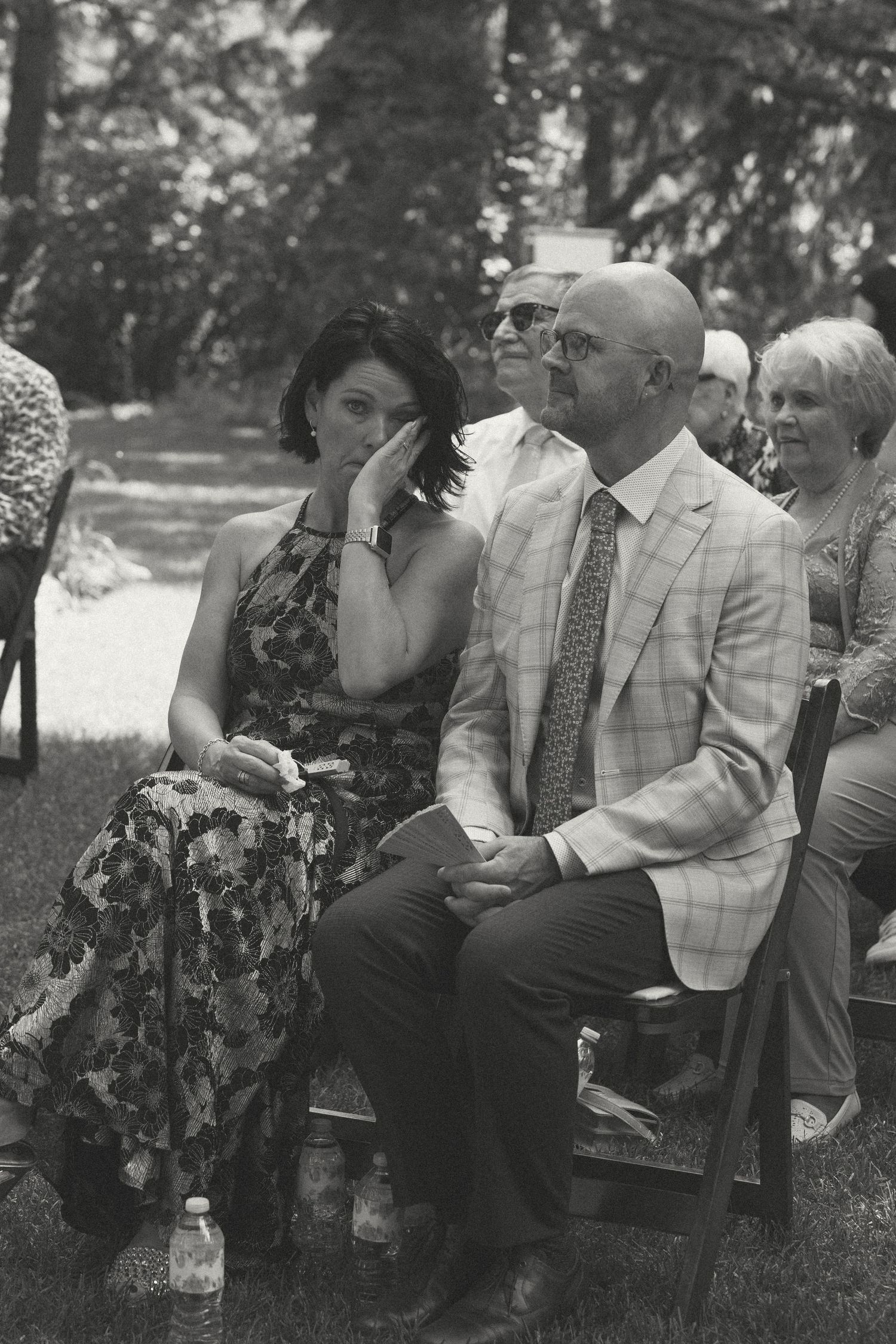 Wedding guests in summer attire seated outdoors watching an emotional ceremony in a garden setting.