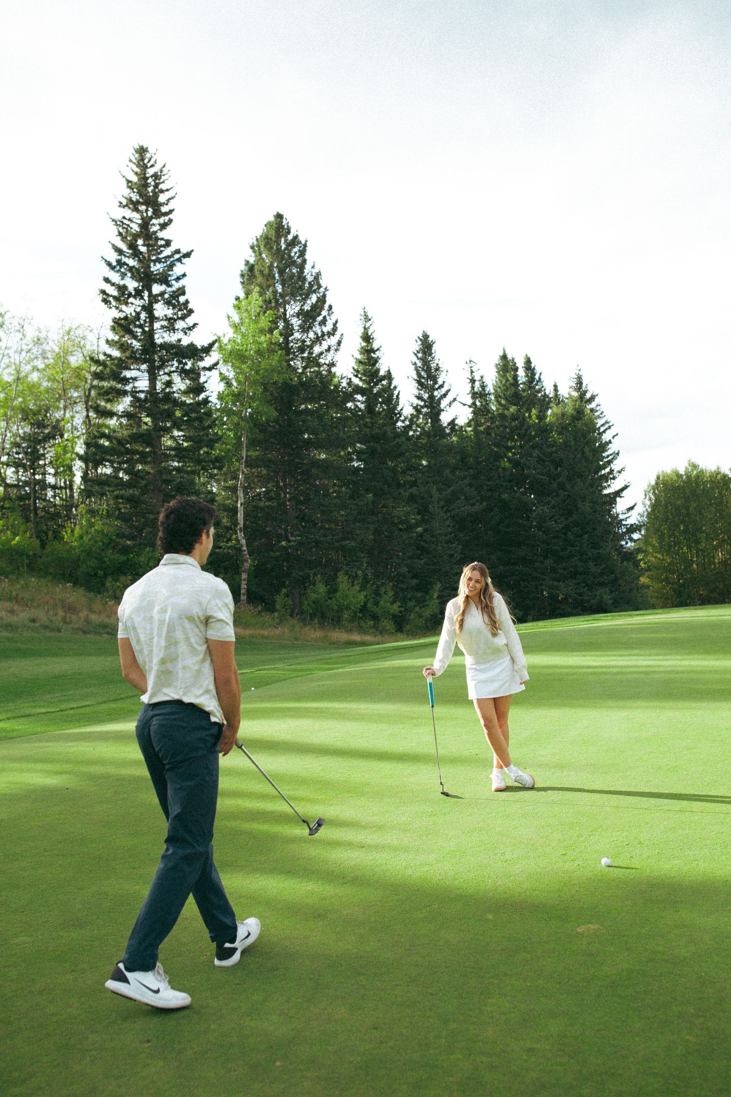 Couple golfing together on manicured green surrounded by evergreen trees.