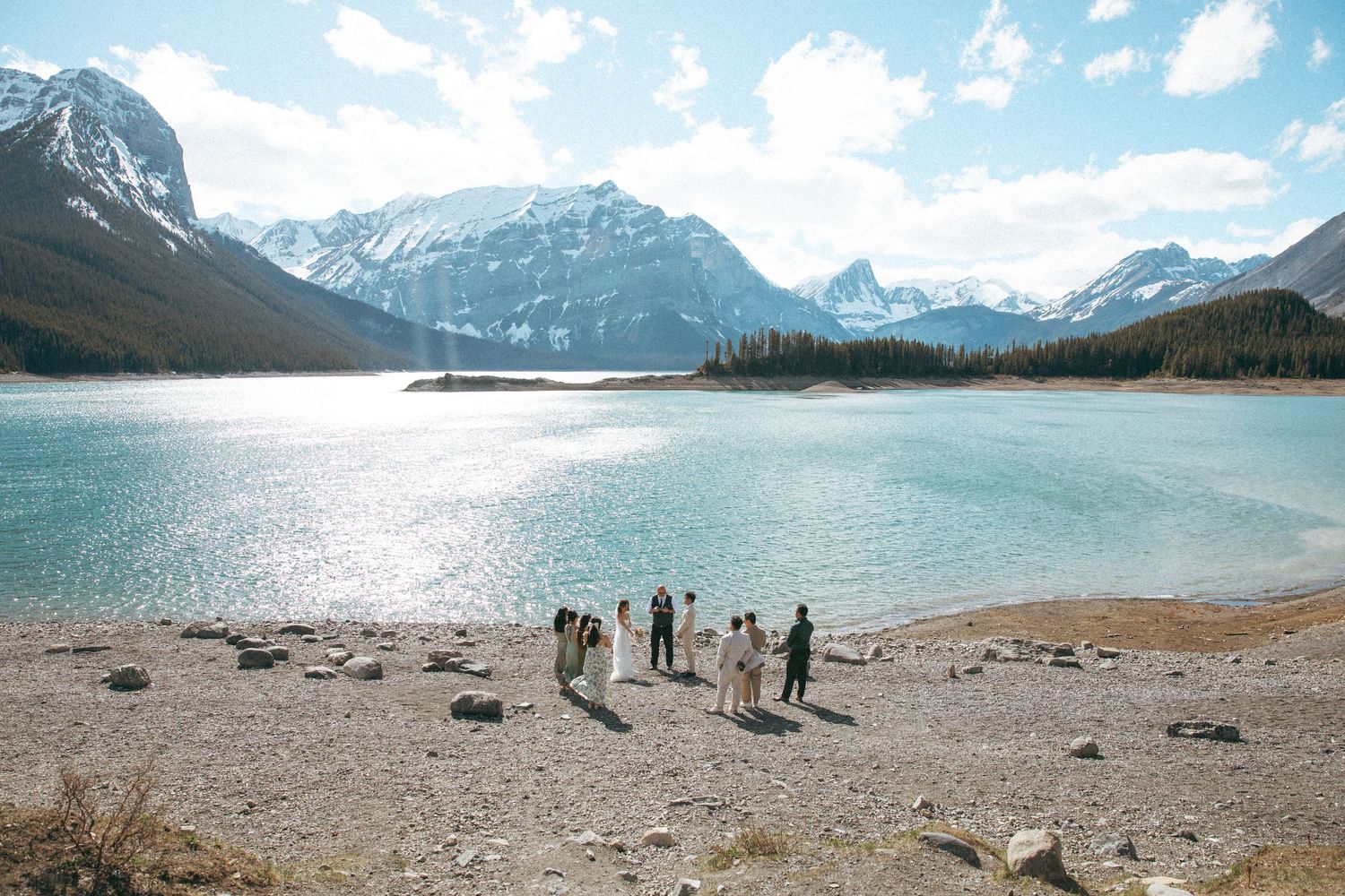 Wedding party poses by a pristine mountain lake with snow-capped peaks in the background.