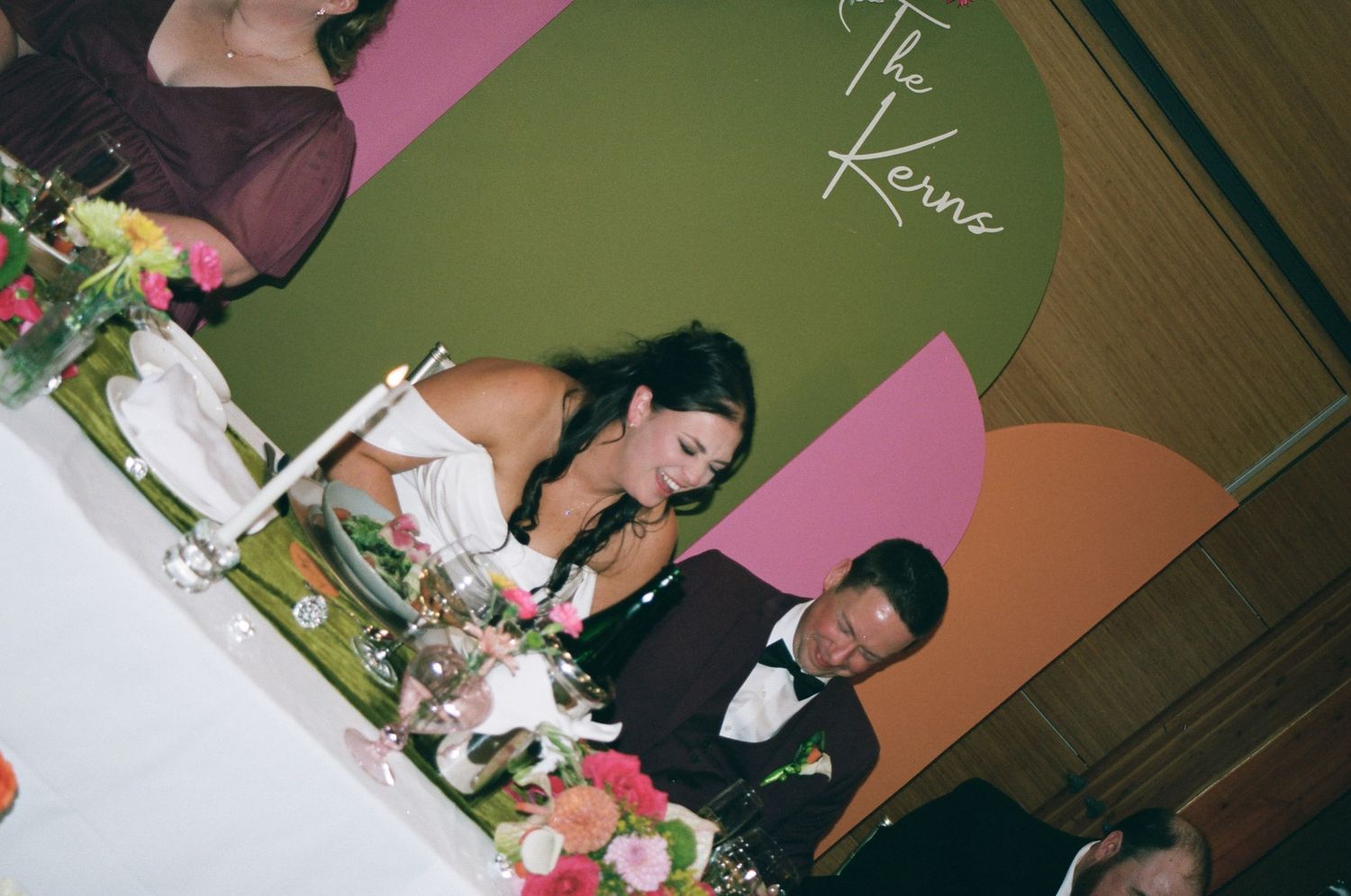 Bride and groom seated at sweetheart table with floral arrangements at wedding reception with custom backdrop.
