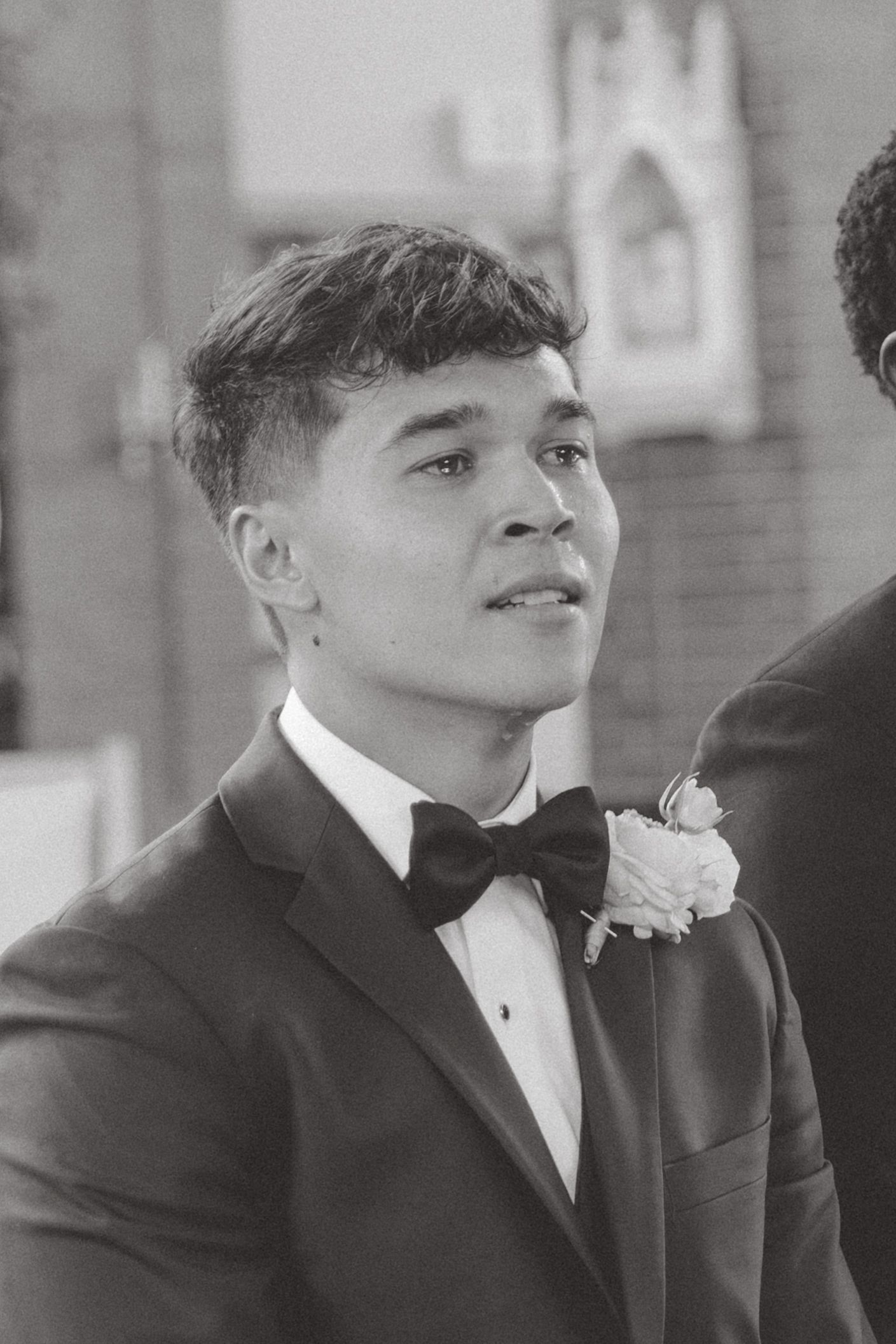Young man in tuxedo with bow tie and boutonniere at formal event, black and white portrait photography.