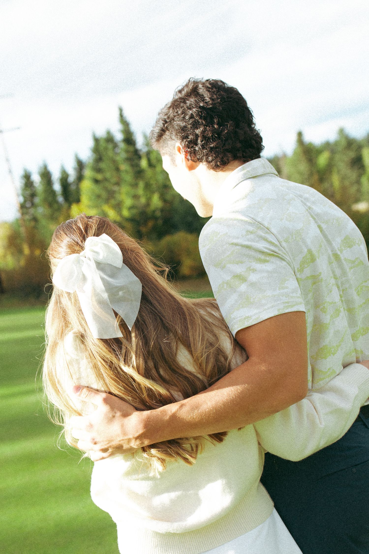Couple embracing on a sunny golf course with pine trees in the background.