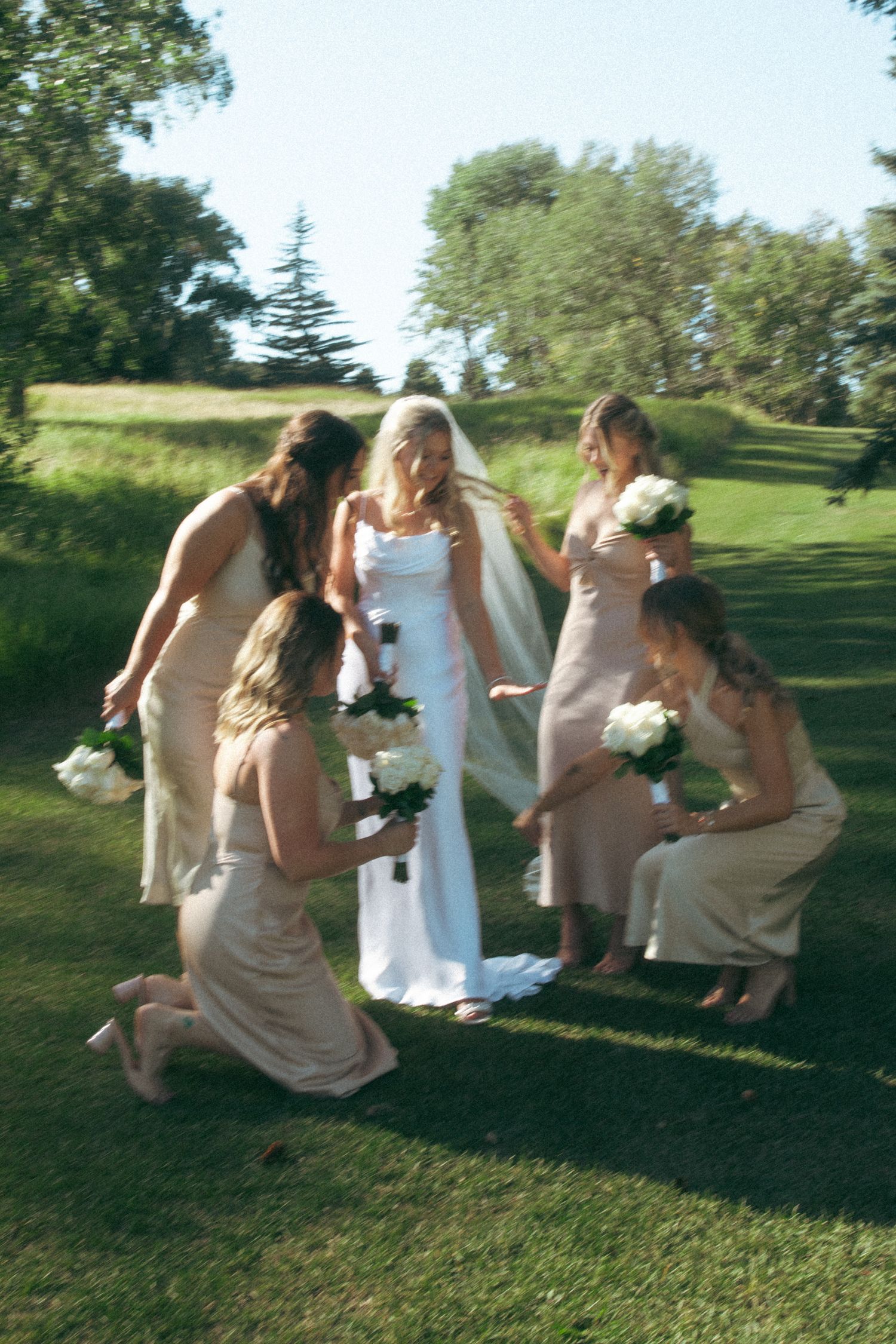 Bridesmaids in champagne dresses gather around the bride in white for a candid outdoor photo.