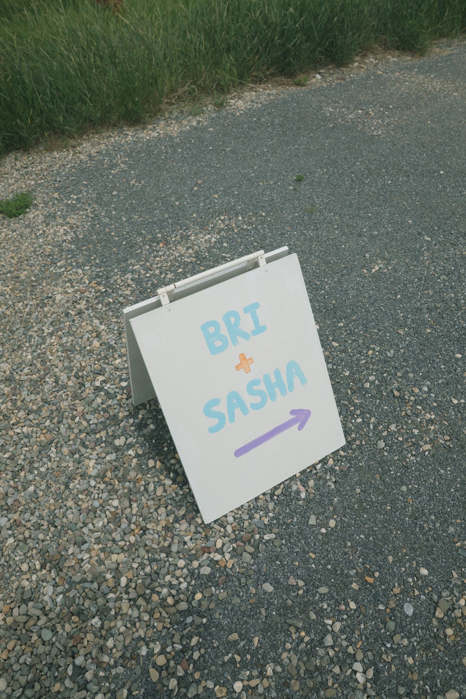A white folded sign with 'BRI + SASHA' written in blue text sits on asphalt pavement.