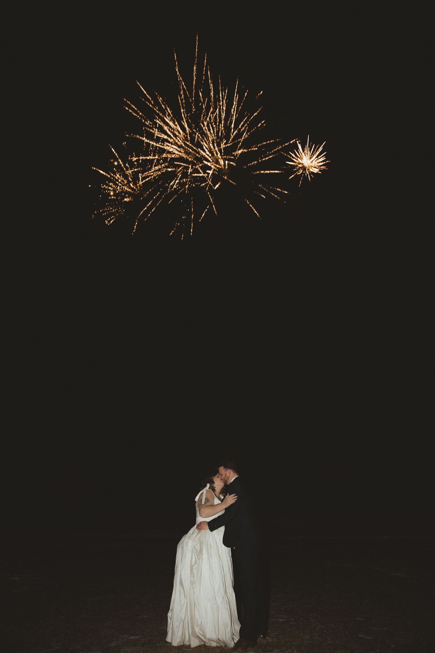 Bride and groom embracing under golden fireworks bursting in dark night sky at wedding celebration.
