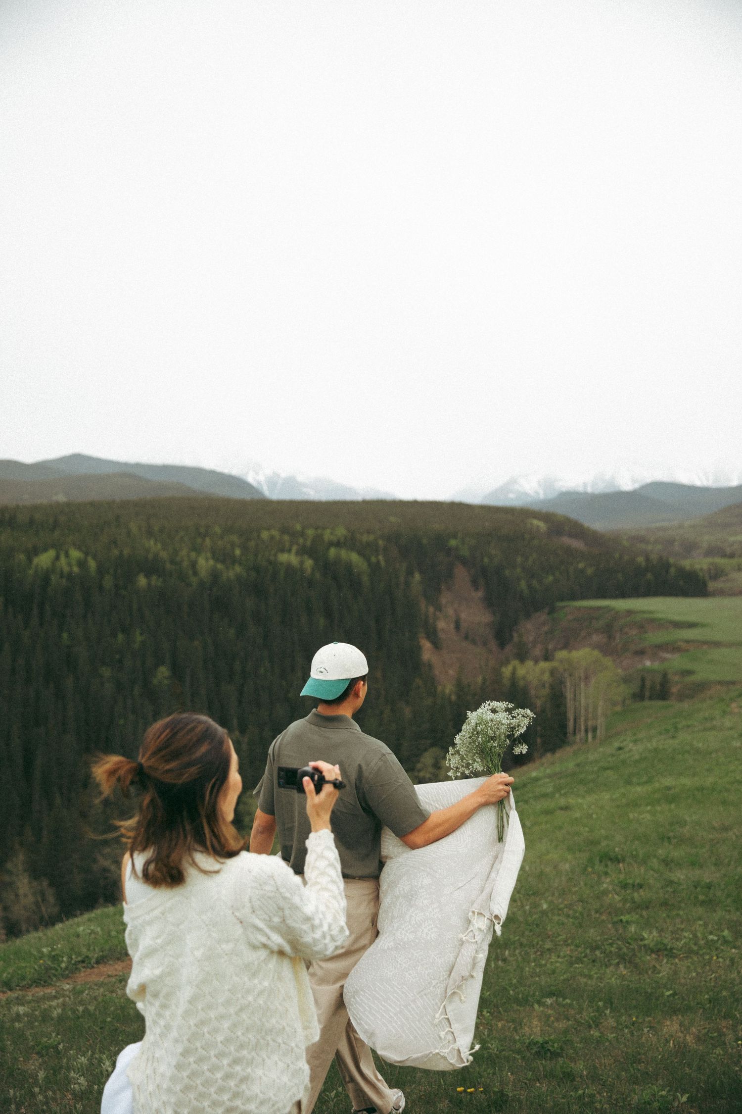 Two people hiking in mountains carry flowers while overlooking forested valley view.
