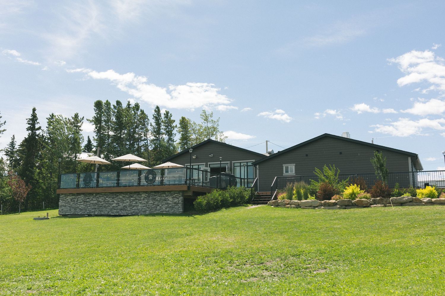 Green countryside lodge with deck and patio umbrellas surrounded by tall trees and manicured lawn on a sunny day.