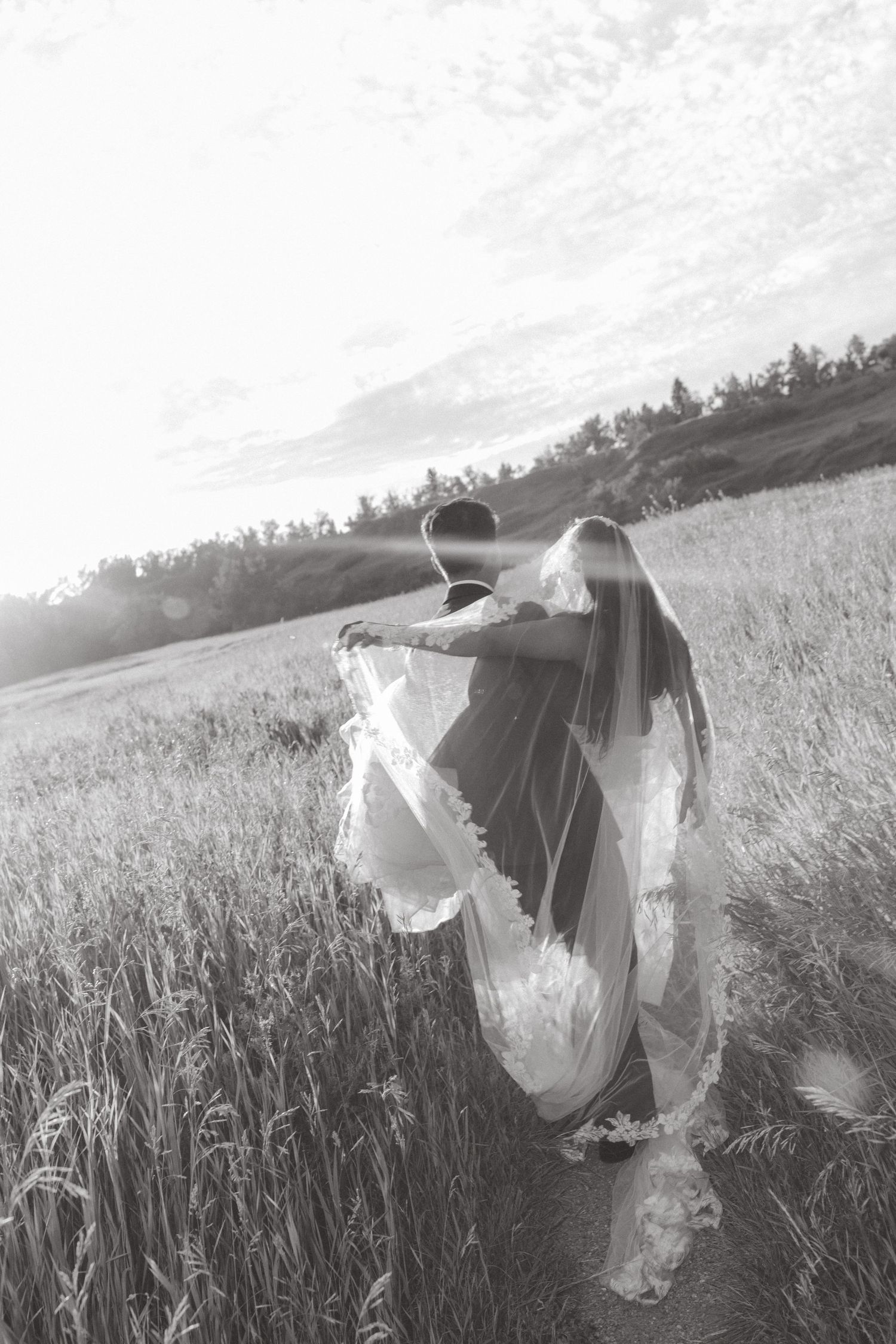 Black and white photo of bride and groom walking through a sunlit field, veil flowing in the breeze.