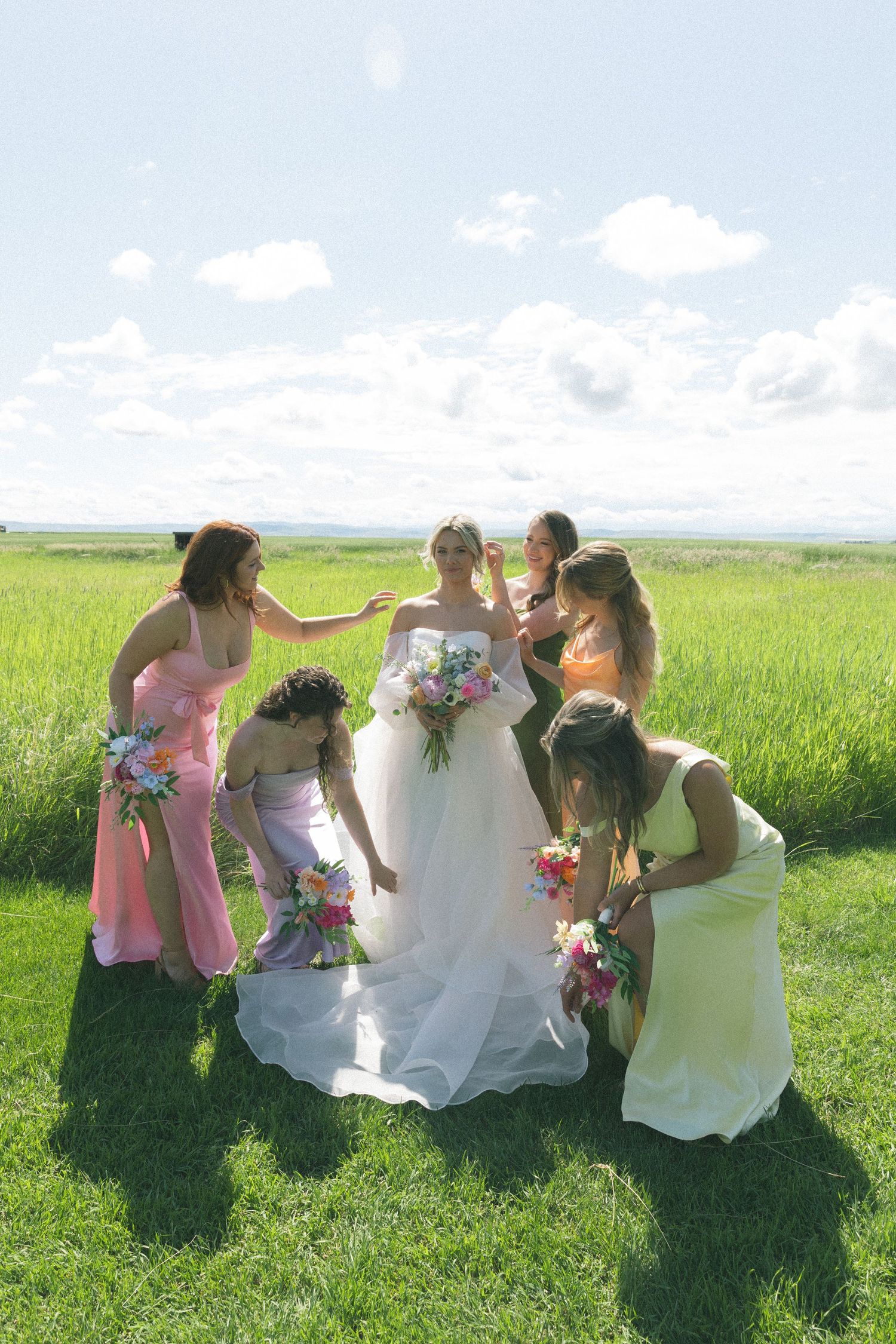 Wedding party members sit together on grass in coordinated dresses against a scenic countryside backdrop.