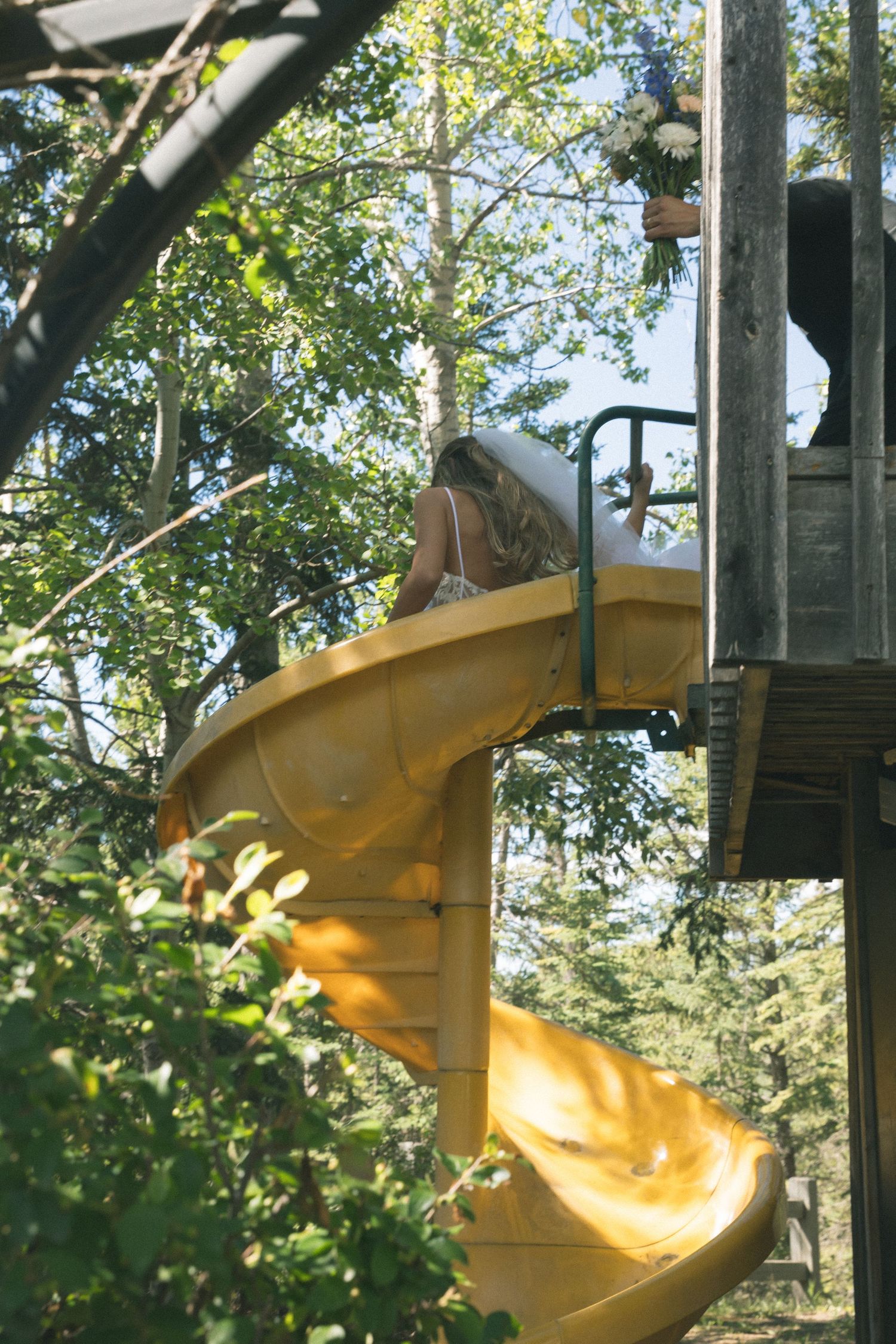 Child climbing a yellow spiral playground slide surrounded by lush green trees outdoors.