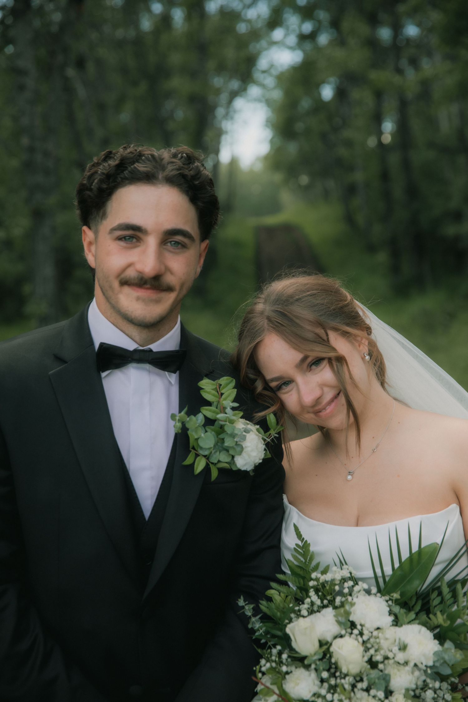 Smiling bride and groom pose outdoors in lush green forest, groom in black tuxedo, bride holding white floral bouquet.