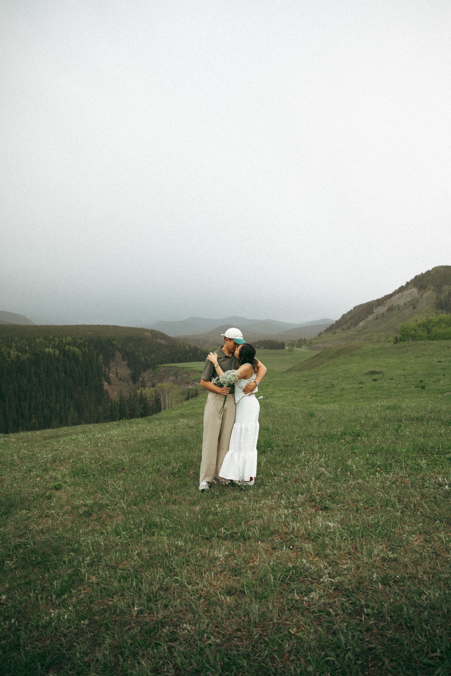 Two people embrace on a grassy hillside overlooking misty mountains on an overcast day.