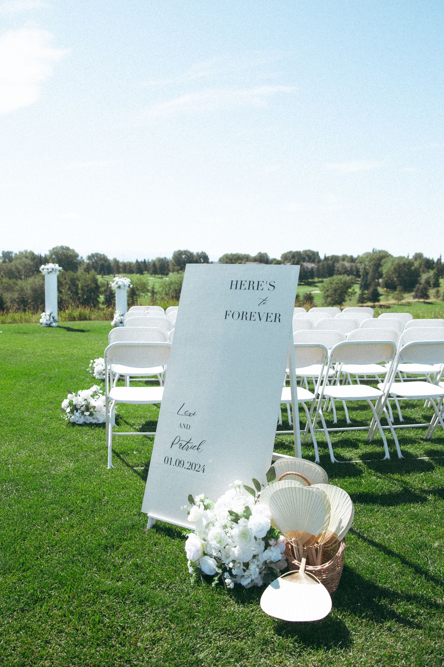 Outdoor wedding ceremony setup with white chairs and 'Here's to Forever' sign on grassy lawn.