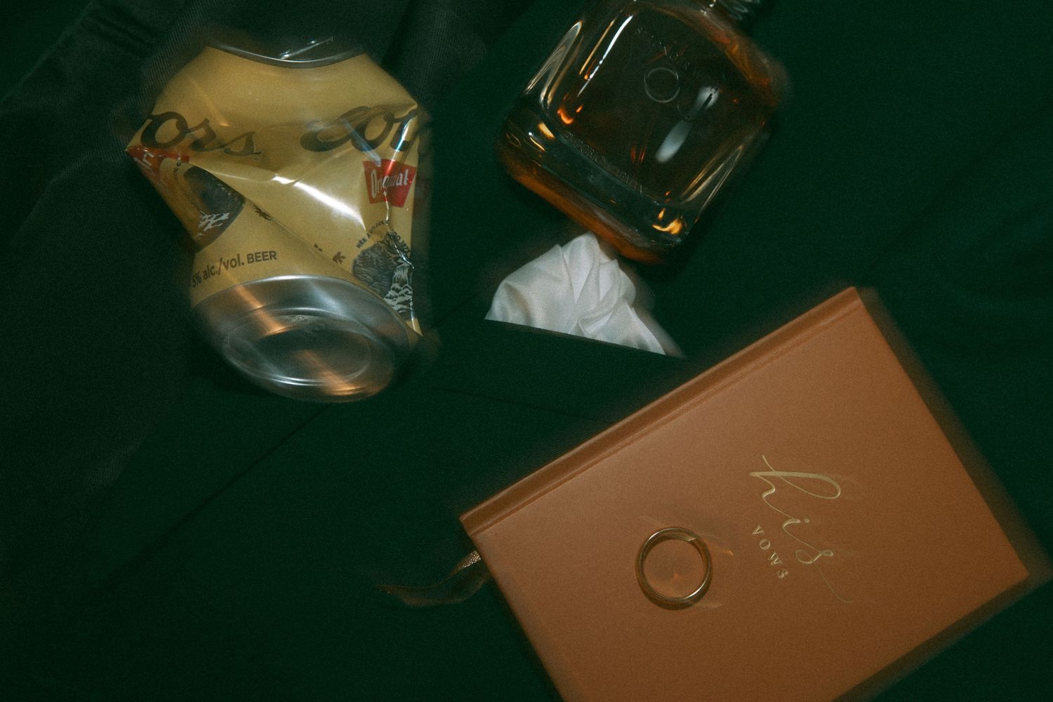 A crushed aluminum can lies next to a glass and brown leather notebook on a dark green surface.