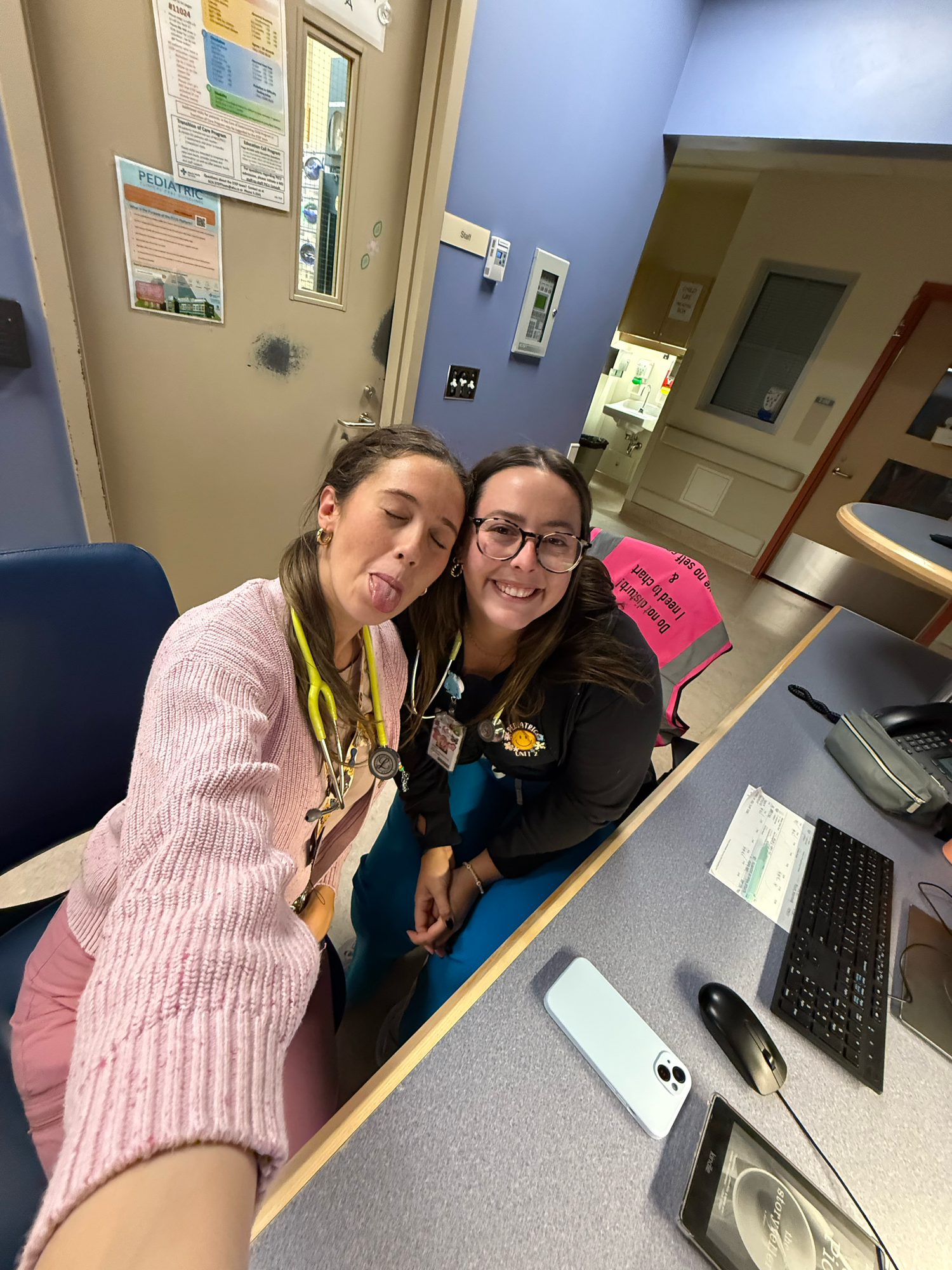 Two healthcare workers taking a playful selfie at a medical station desk.