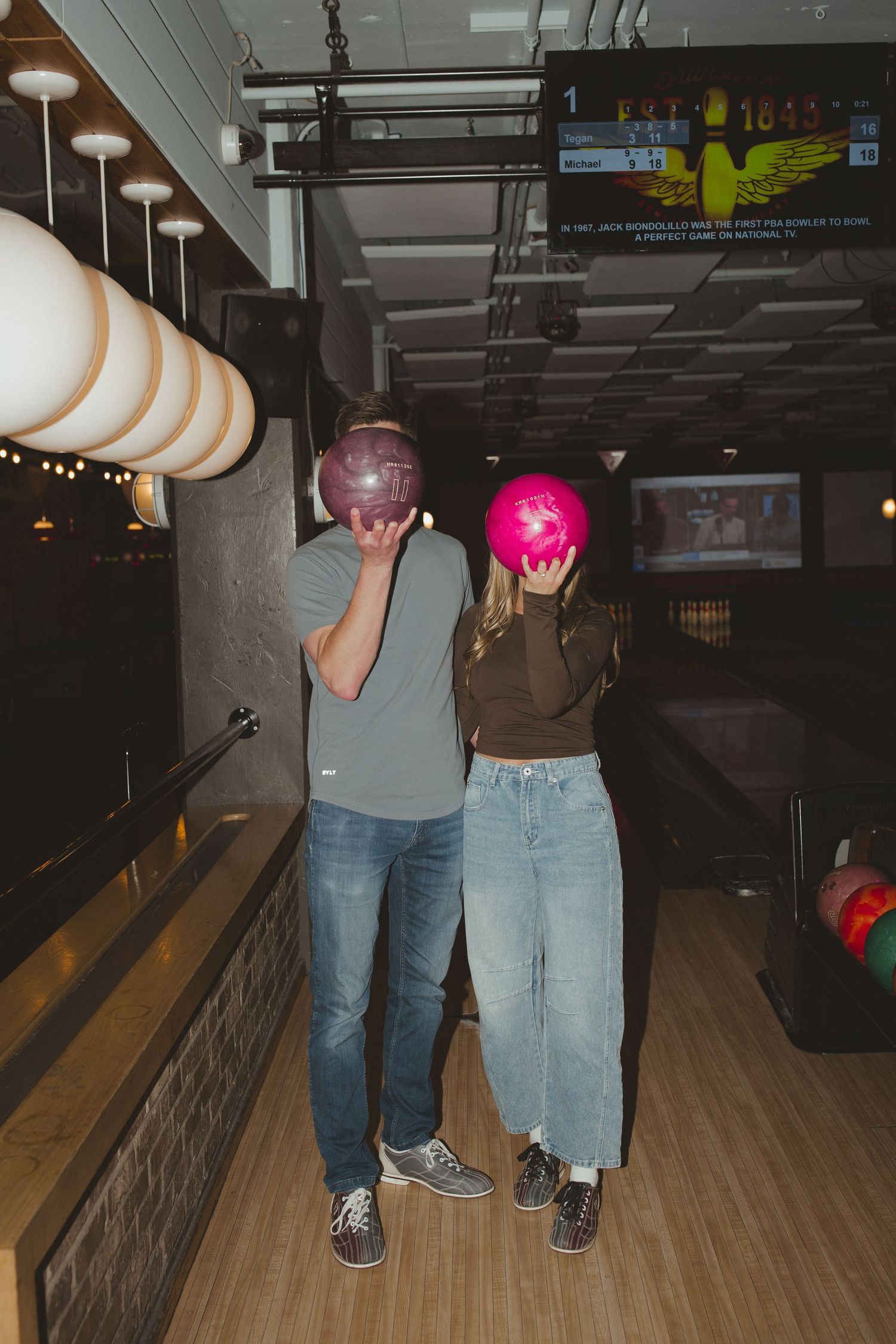 Couple holding colorful bowling balls in front of their faces at a bowling alley lane.