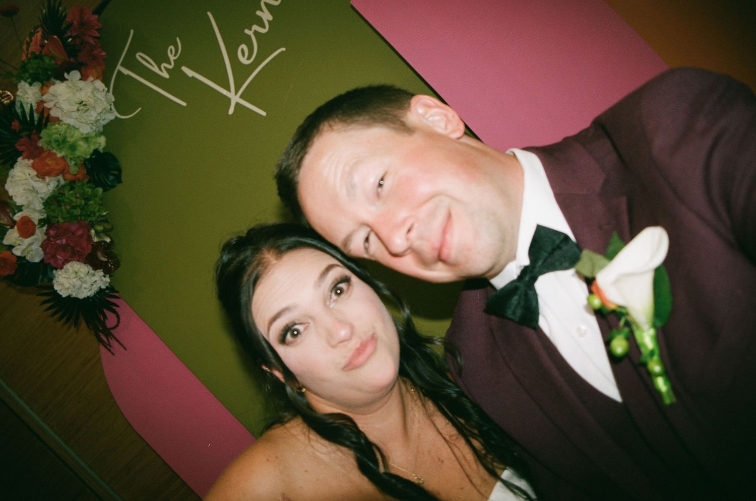 A couple posing together at a wedding reception, man in burgundy tuxedo with boutonniere, woman in white dress.