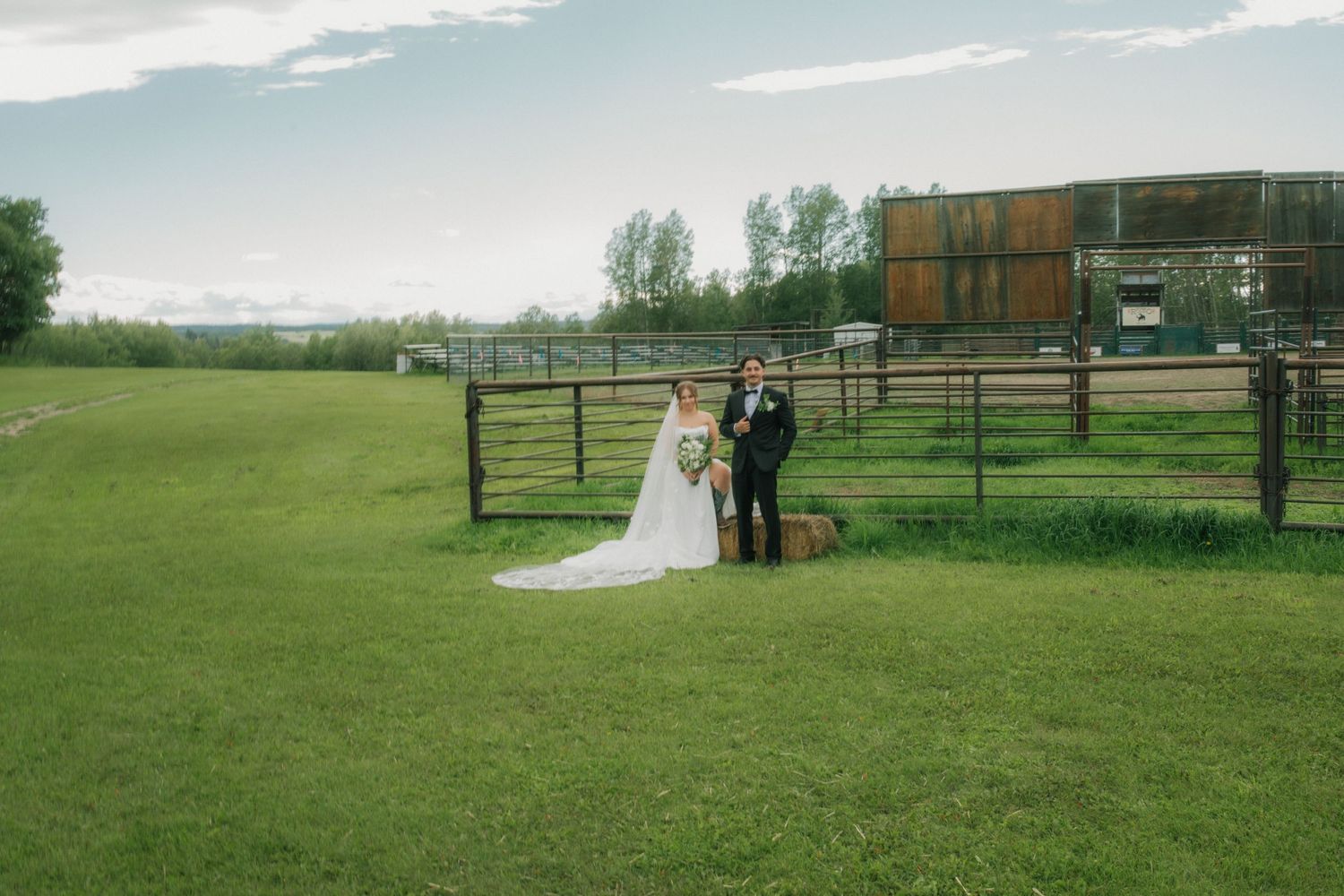 Bride and groom standing together on green grass near a rustic farm fence on their wedding day.