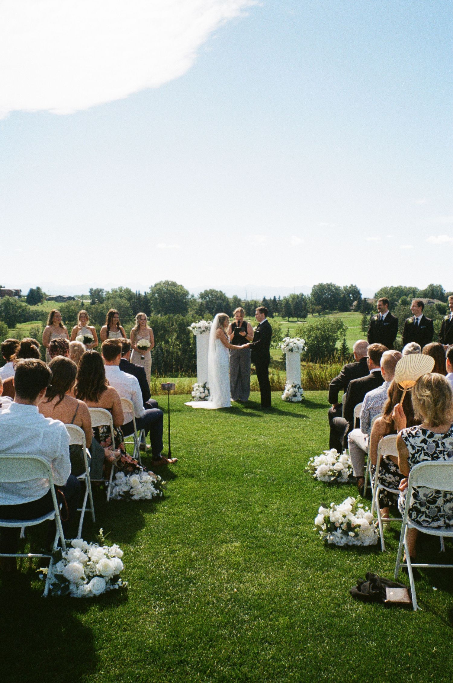 Outdoor wedding ceremony on a lush green hillside with white chairs and floral aisle decorations under a bright blue sky.