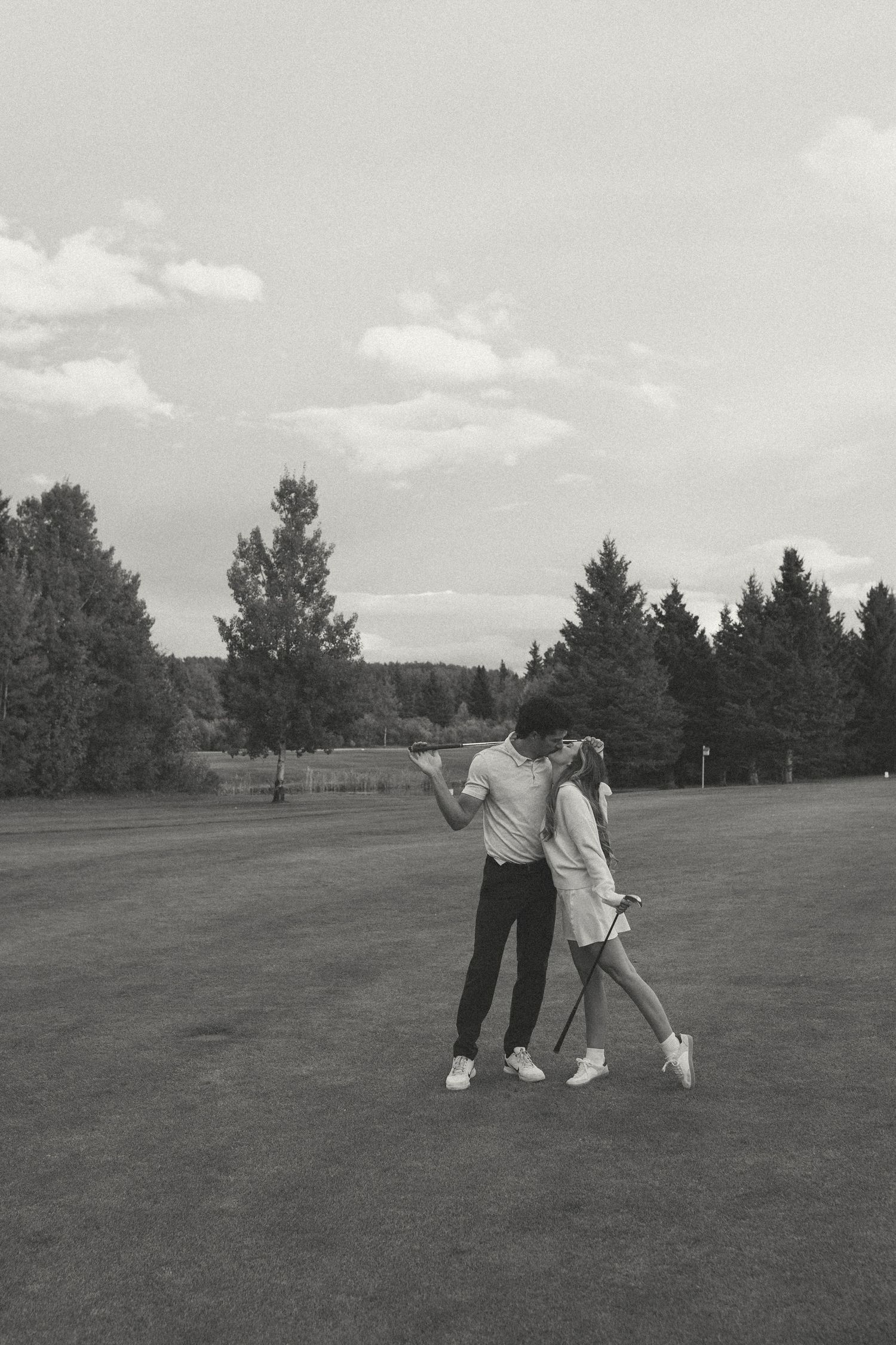 Couple in casual attire posing with golf clubs on course in vintage black and white style.