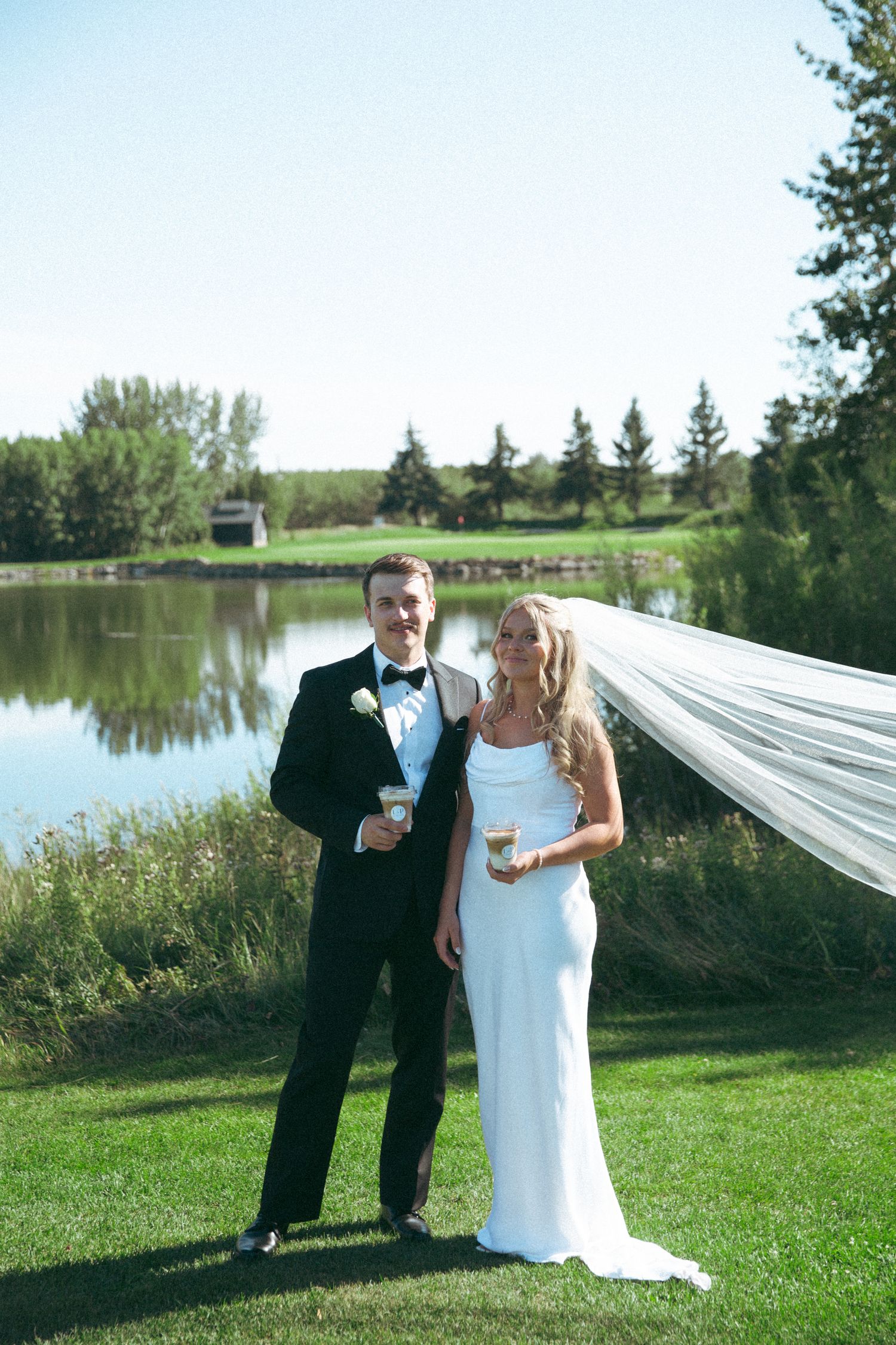 Newlyweds pose together by a pond with flowing veil and formal attire on their wedding day.