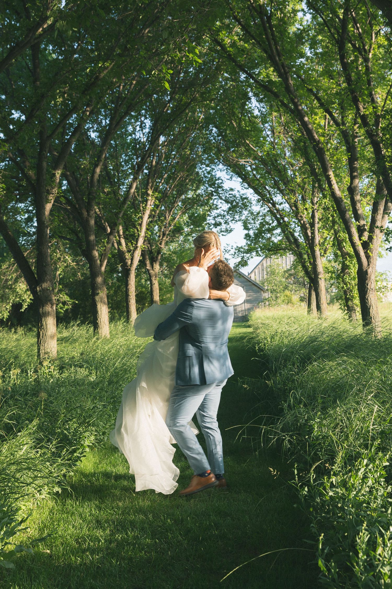 Wedding couple in formal attire playfully running and dancing through a tree-lined path in a park.