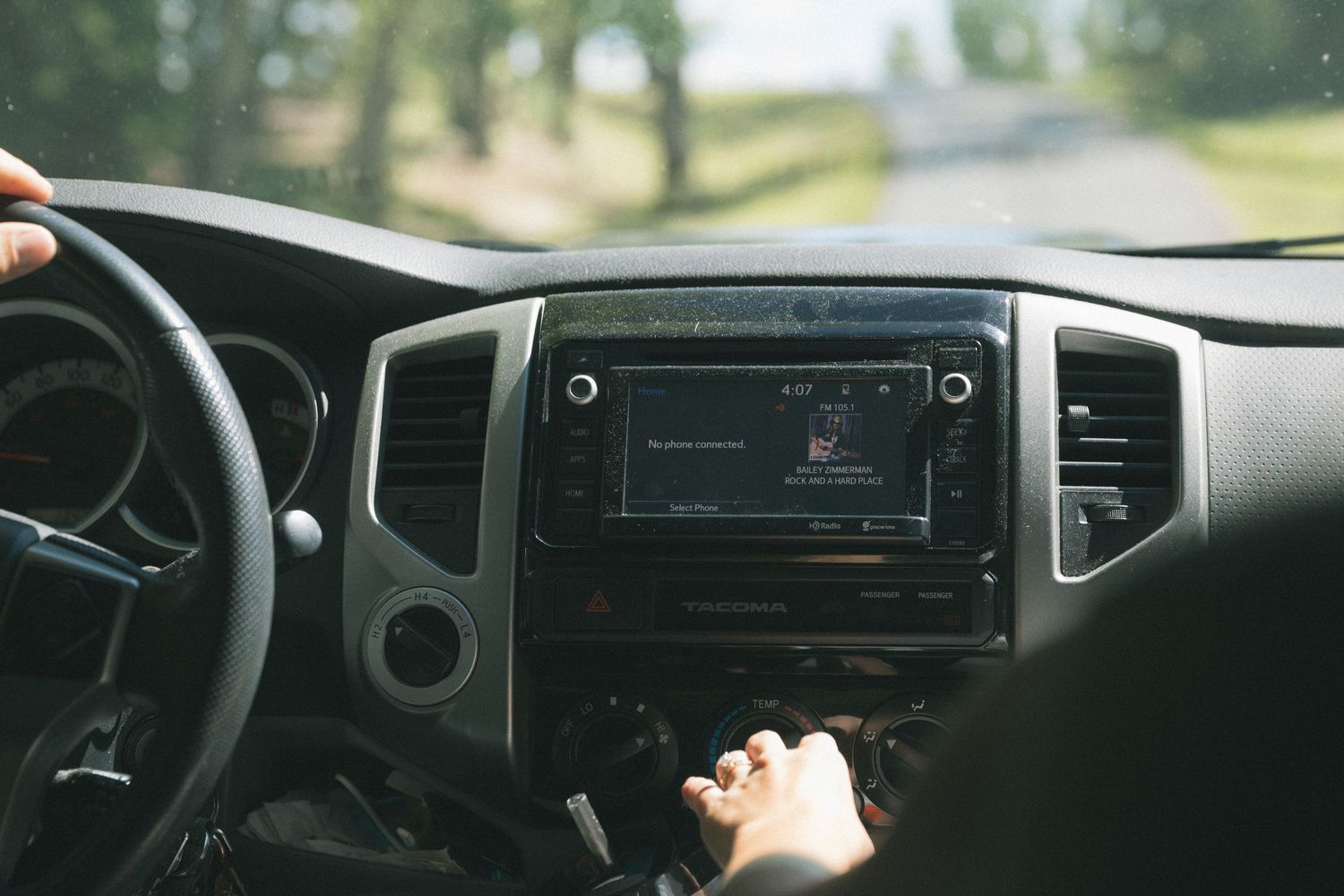 Interior view of a car dashboard with a touchscreen radio, steering wheel, and hands on the console while driving.