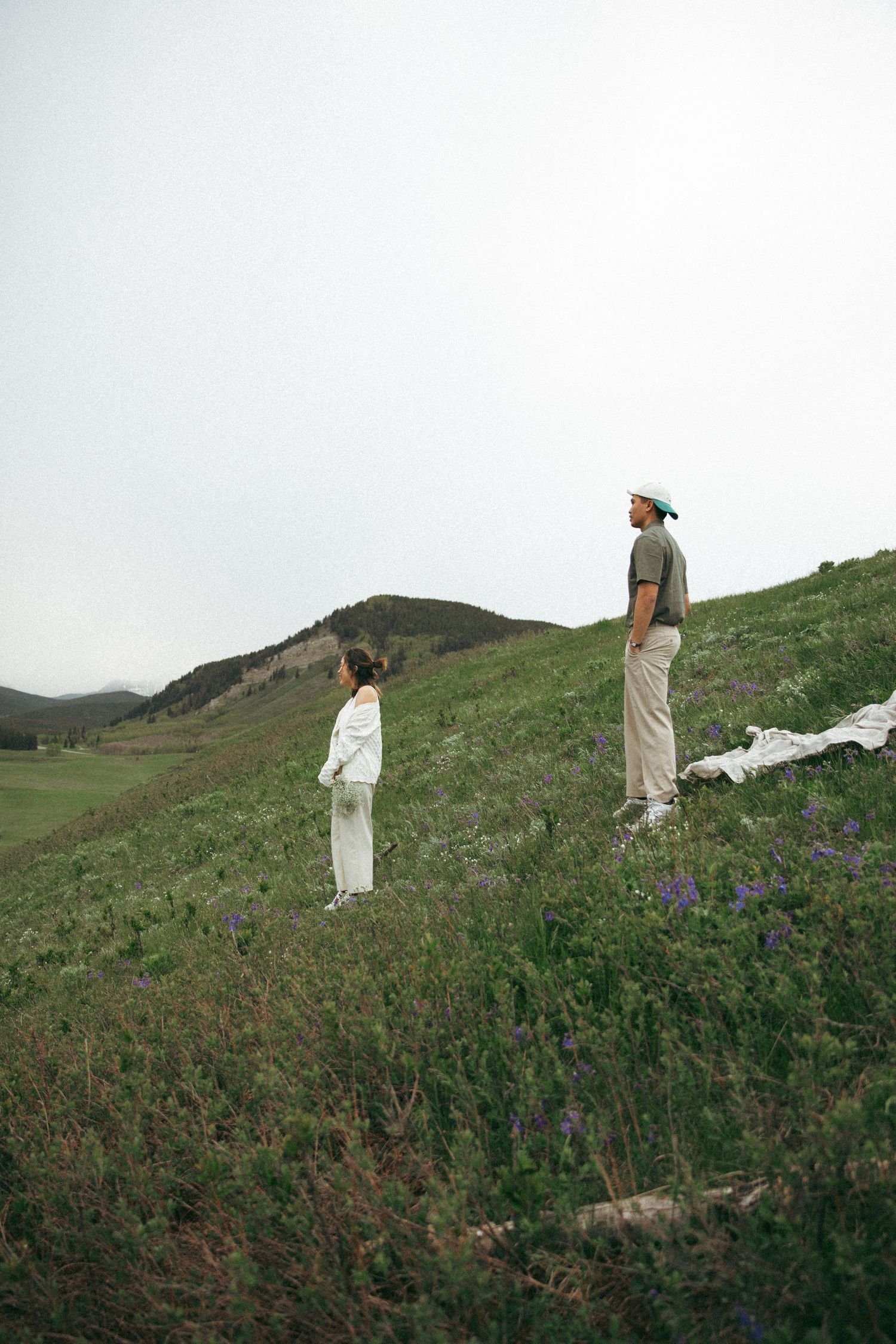 Two people standing on a grassy hillside with mountains in the distance.