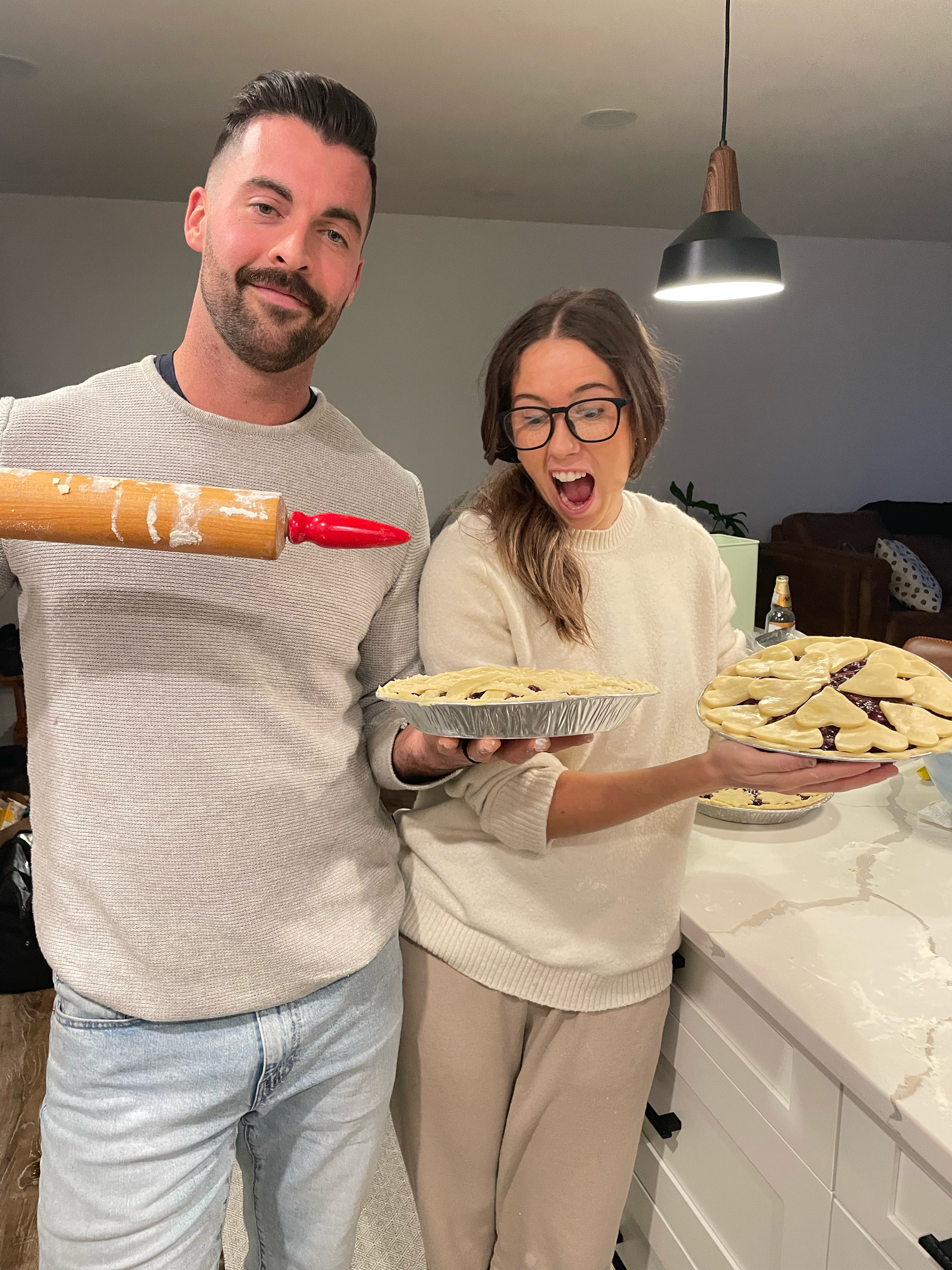 Two people in kitchen with baking supplies and pies showing excited expressions.