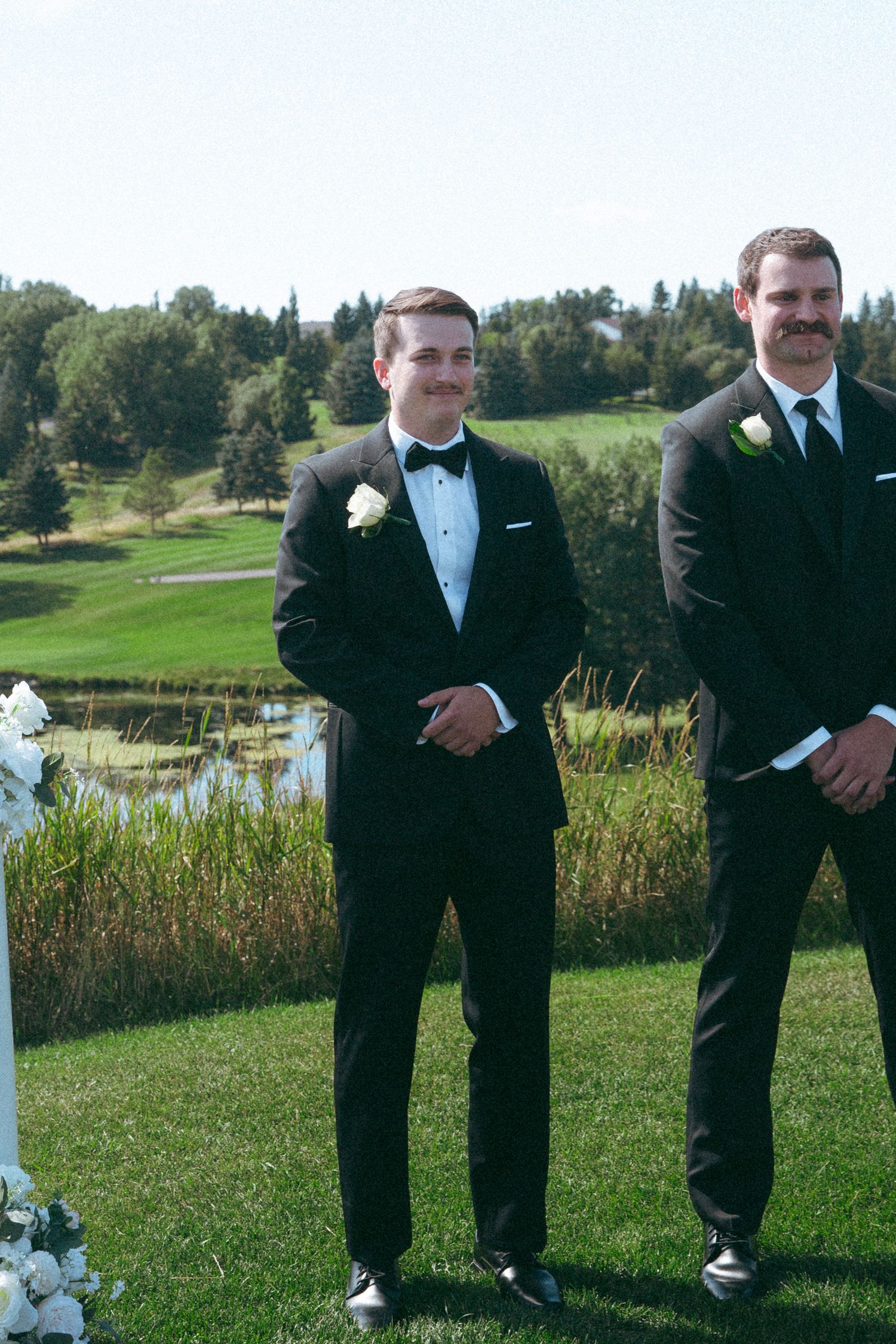Groomsmen in black tuxedos stand at outdoor wedding ceremony with pond in background.