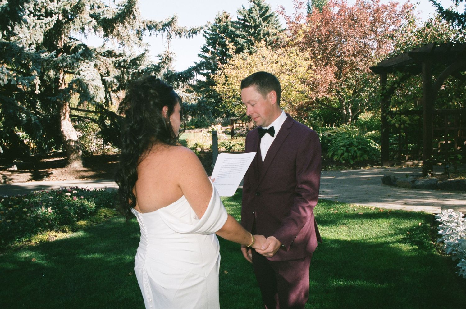 Couple exchanging vows outdoors in a lush garden, groom in burgundy tuxedo holding paper, bride in white strapless gown.