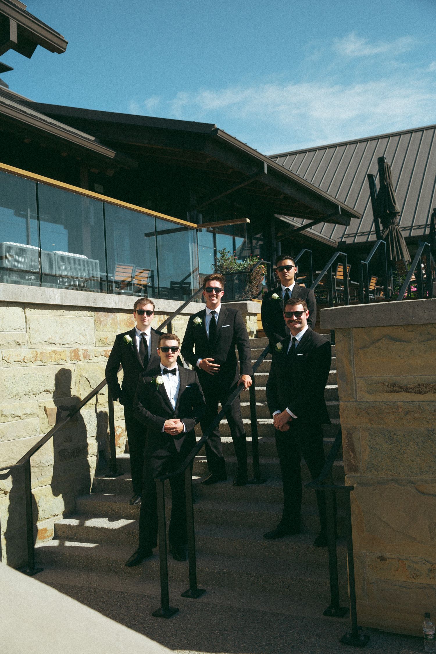 Groomsmen in formal suits and sunglasses posing on stone steps outdoors.