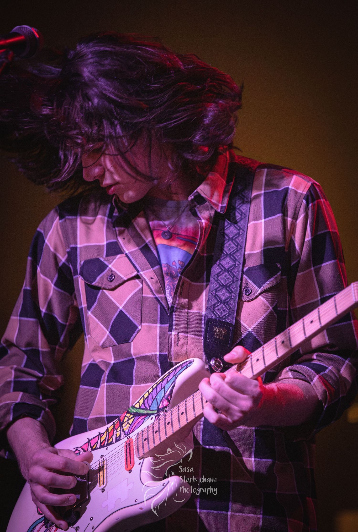 A guitarist in a pink plaid shirt plays passionately under purple stage lighting.