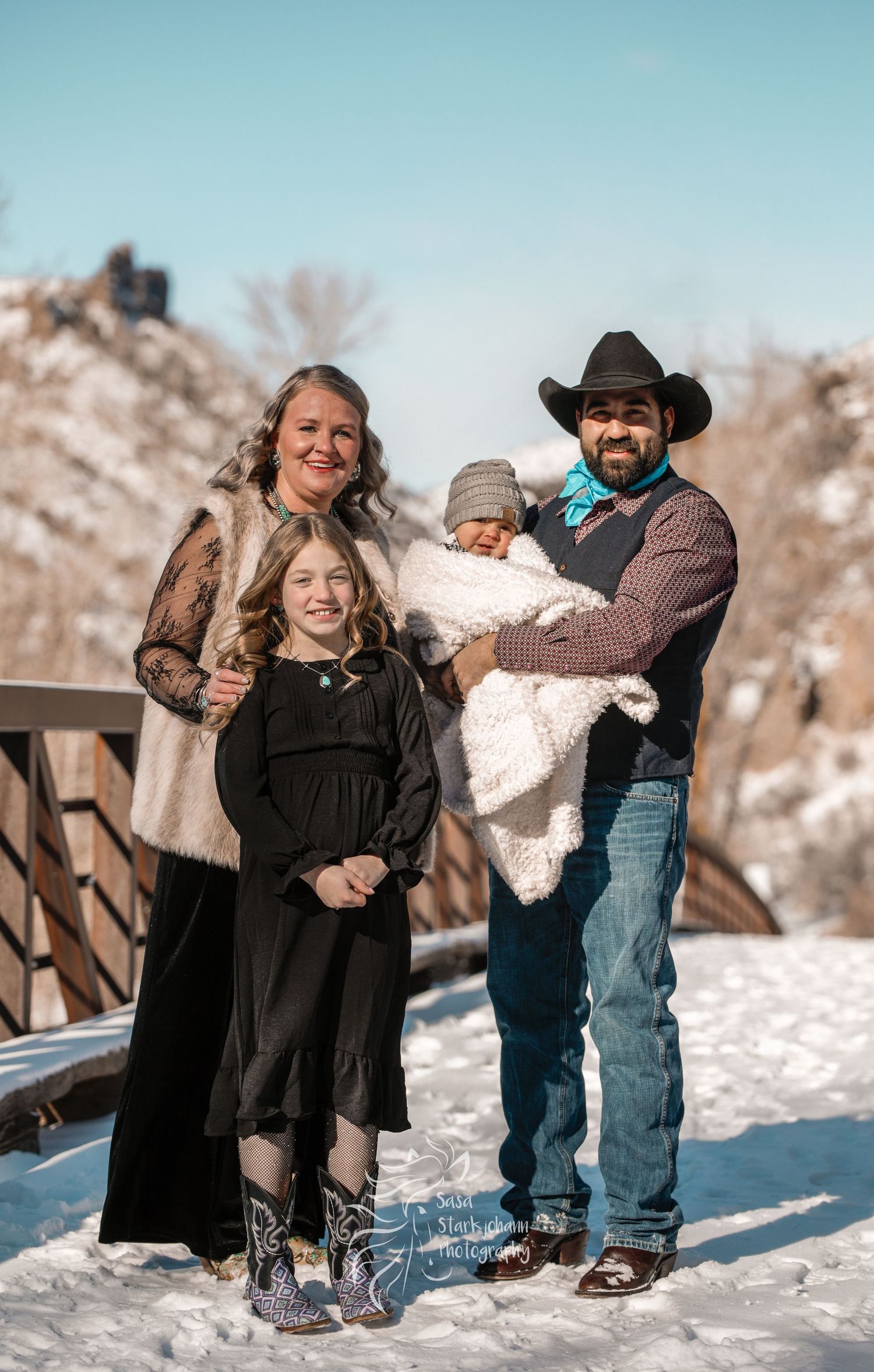 A family stands together on a bridge during winter with snow-capped mountains in the background.