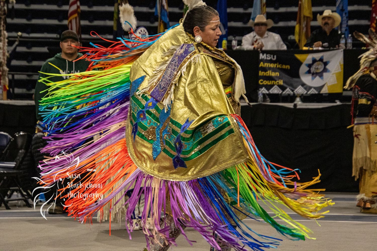 Dancer in golden shawl with rainbow fringe performing traditional dance movements at indoor event.