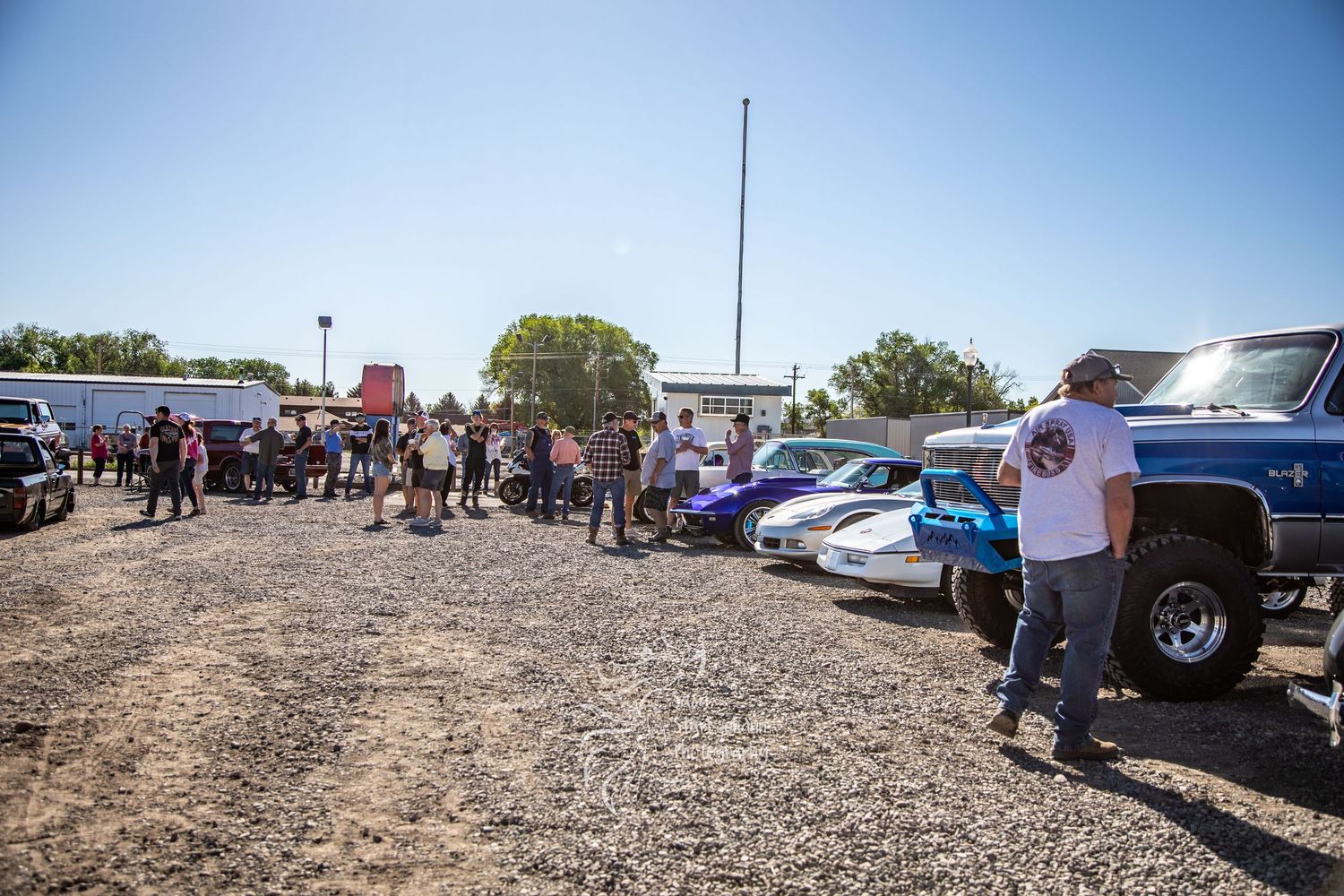A crowd gathers around parked vehicles at a rural community car meet on dirt ground.
