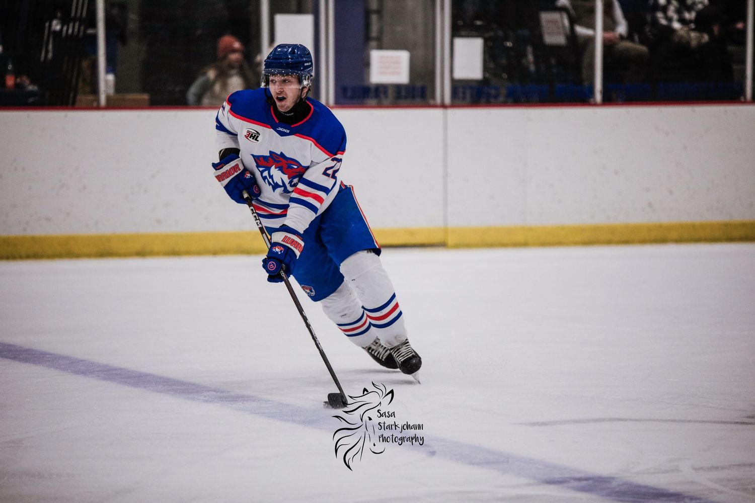 A hockey player in blue and white jersey skates with the puck on an indoor ice rink.