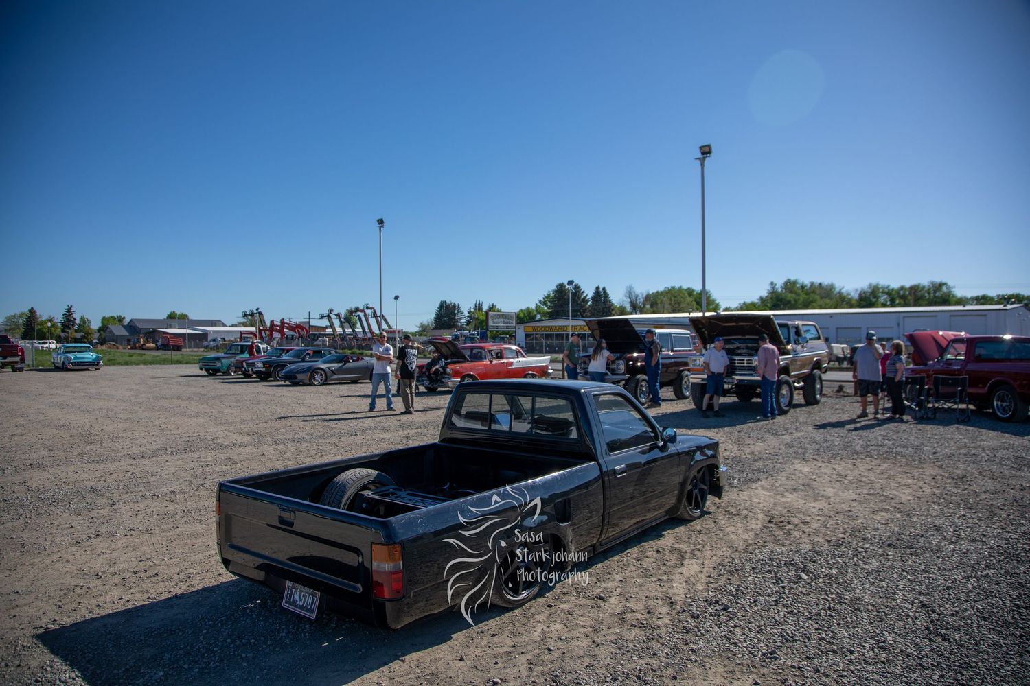 A lowered black pickup truck parked at an outdoor event with other vehicles and spectators visible in bright sunlight.